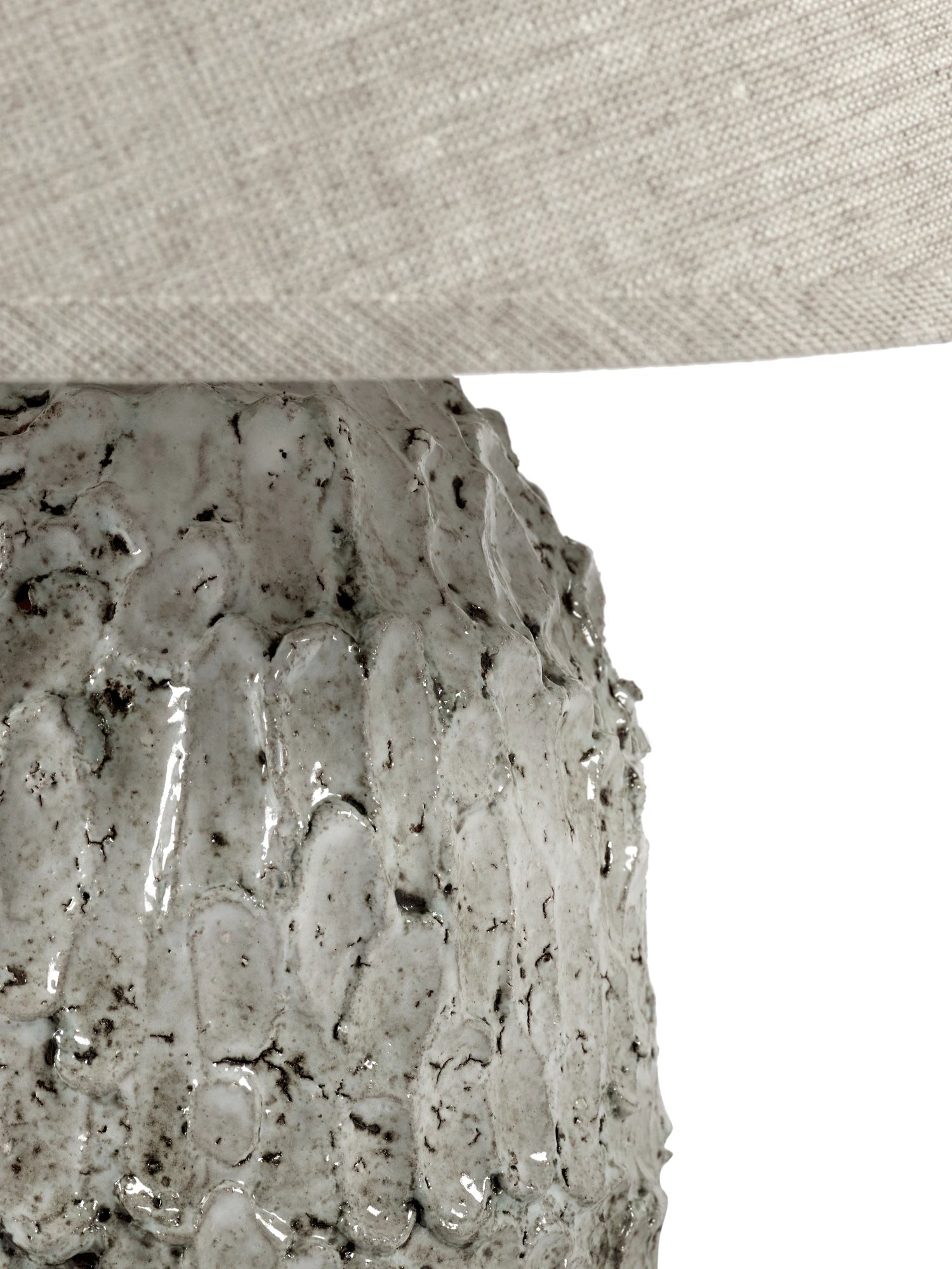 A close-up of the small Tronco ceramic table lamp's base, focusing on the highly textured, glossy light beige/off-white finish with dark speckles, which resembles bark or lava. The lower edge of the natural linen lampshade is visible at the top. The image is cropped to highlight the unique texture of the base.
