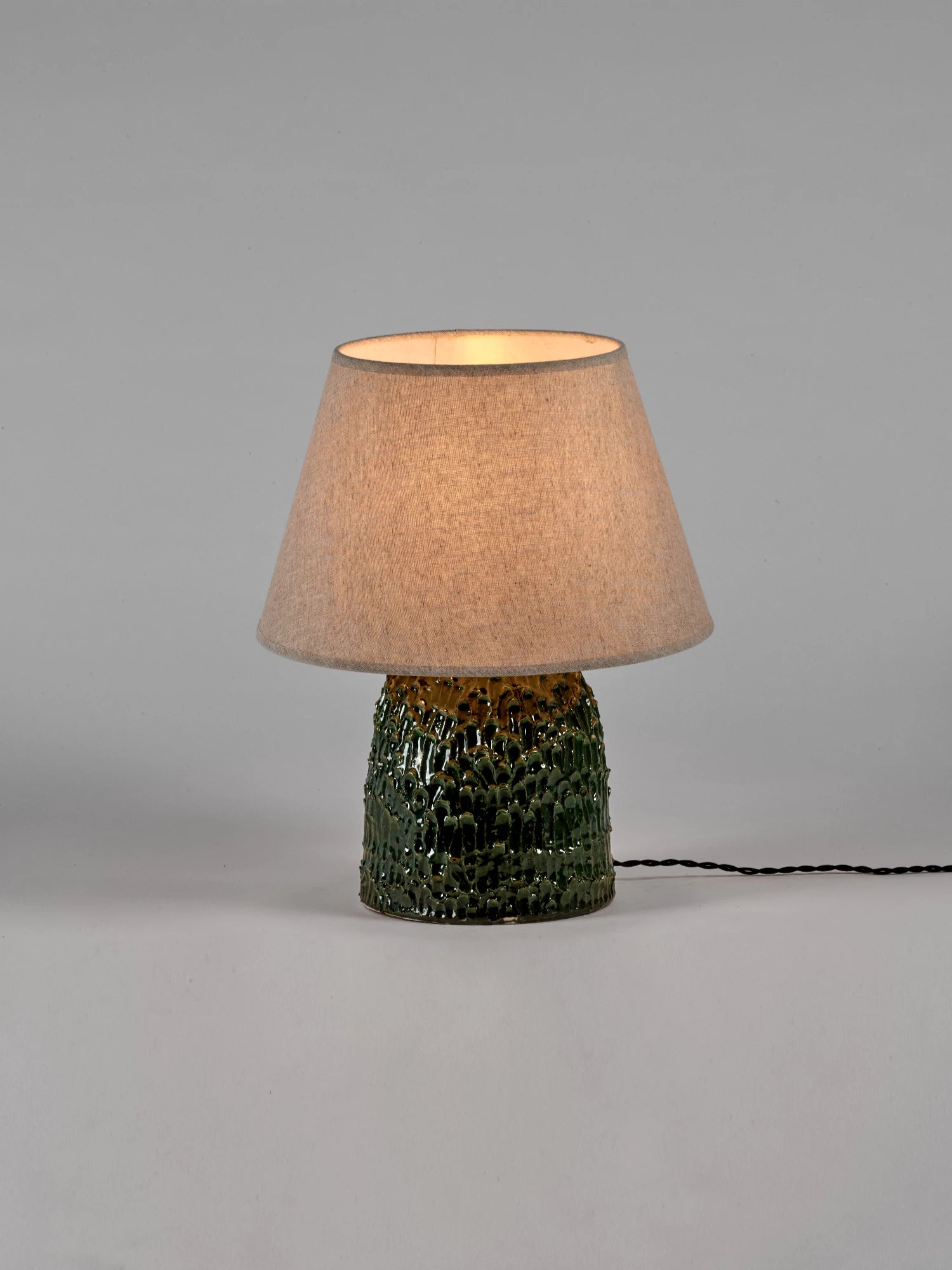 A medium-sized Tronco ceramic table lamp with a dark green, highly textured, glossy base and a conical, natural linen-colored lampshade. The light is switched on, giving the shade a warm glow. The lamp is photographed against a neutral gray background with a black electrical cord extending to the right.