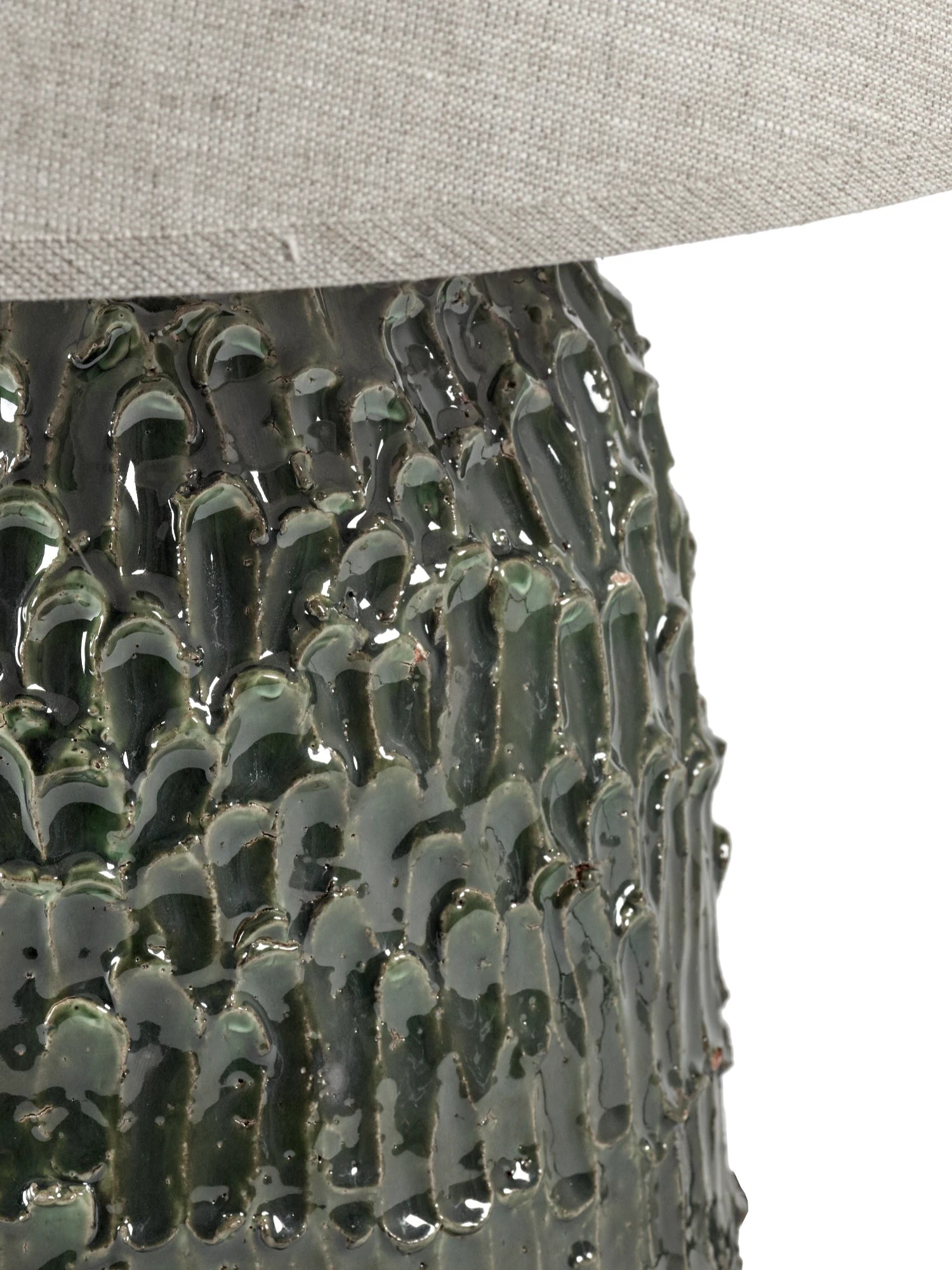 A close-up of the Tronco ceramic table lamp's base, focusing on the highly textured, glossy dark green finish that resembles bark or lava. The lower edge of the neutral white/beige fabric lampshade is visible at the top. The image is cropped to highlight the texture of the base.