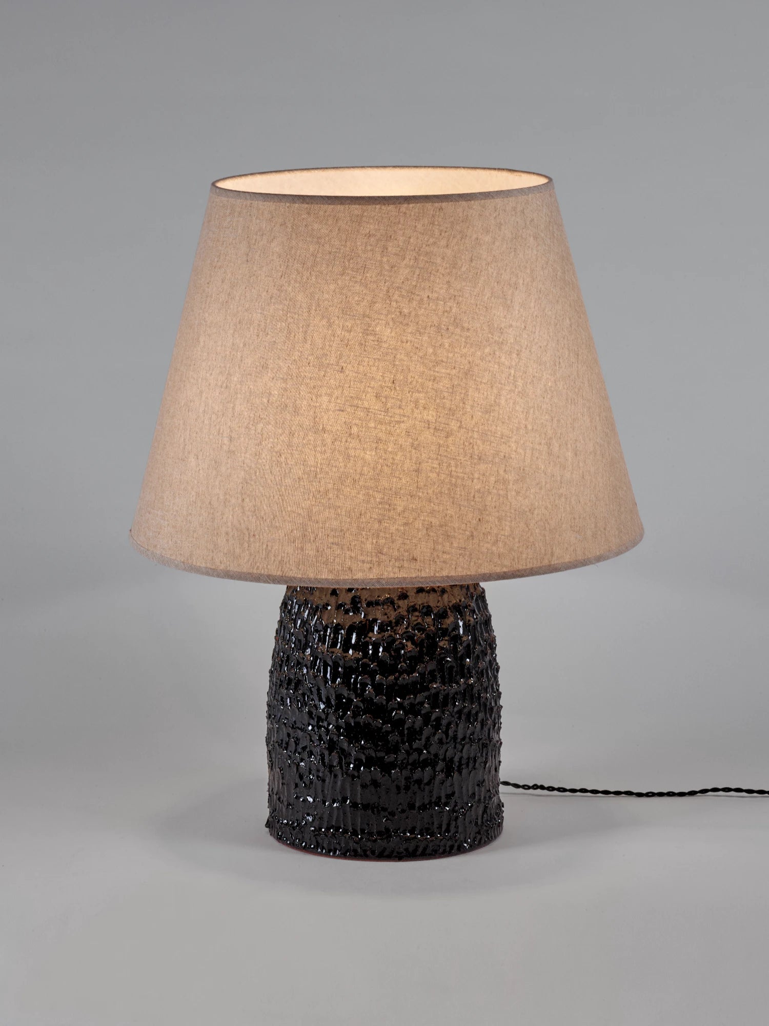 Black Tronco ceramic table lamp with a highly textured, glossy, dark brown/black base and a large, conical, natural linen-colored lampshade. The lamp is photographed against a neutral gray background. A black electrical cord extends to the right.
