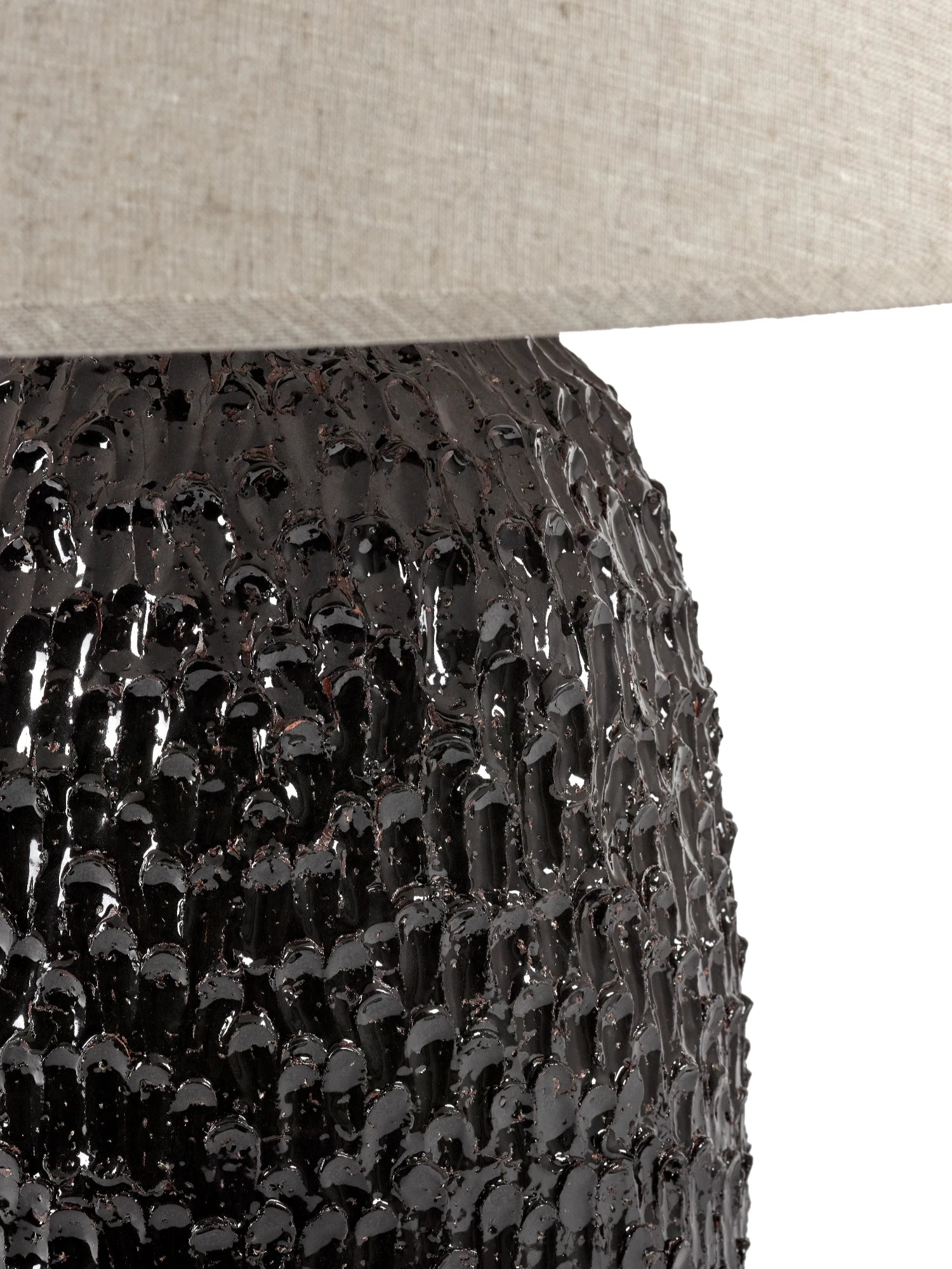 A close-up of the Tronco ceramic table lamp's base, showing the highly textured, glossy dark brown/black finish that resembles bark or lava. The bottom edge of the natural linen lampshade is visible at the top. The image is cropped to focus on the texture of the base.