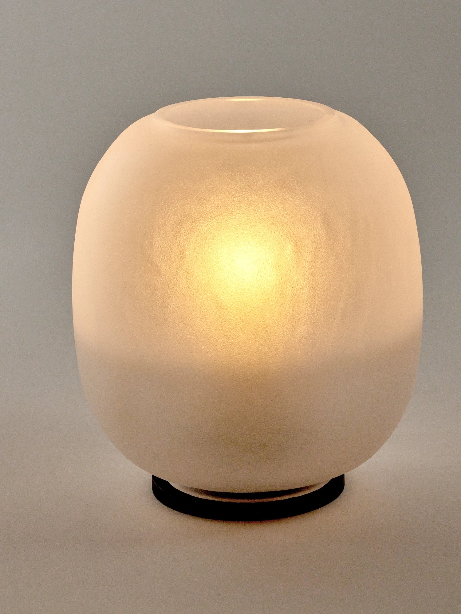 A studio close-up of the Lumen Wind Light, showing its smooth, frosted, matte glass globe resting on a thin, black circular base. The light inside is turned on, casting a bright, warm yellow-orange glow that illuminates the center of the globe and fades toward the top and bottom. The background is a neutral gray.