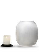 The Lumen Wind Light is shown disassembled on a white background. The frosted, matte glass globe sits on the right, and the small, battery-operated LED pillar candle is placed next to it on the left, resting on the thin black circular base. The candle is lit, showing a warm, flickering glow at the top.