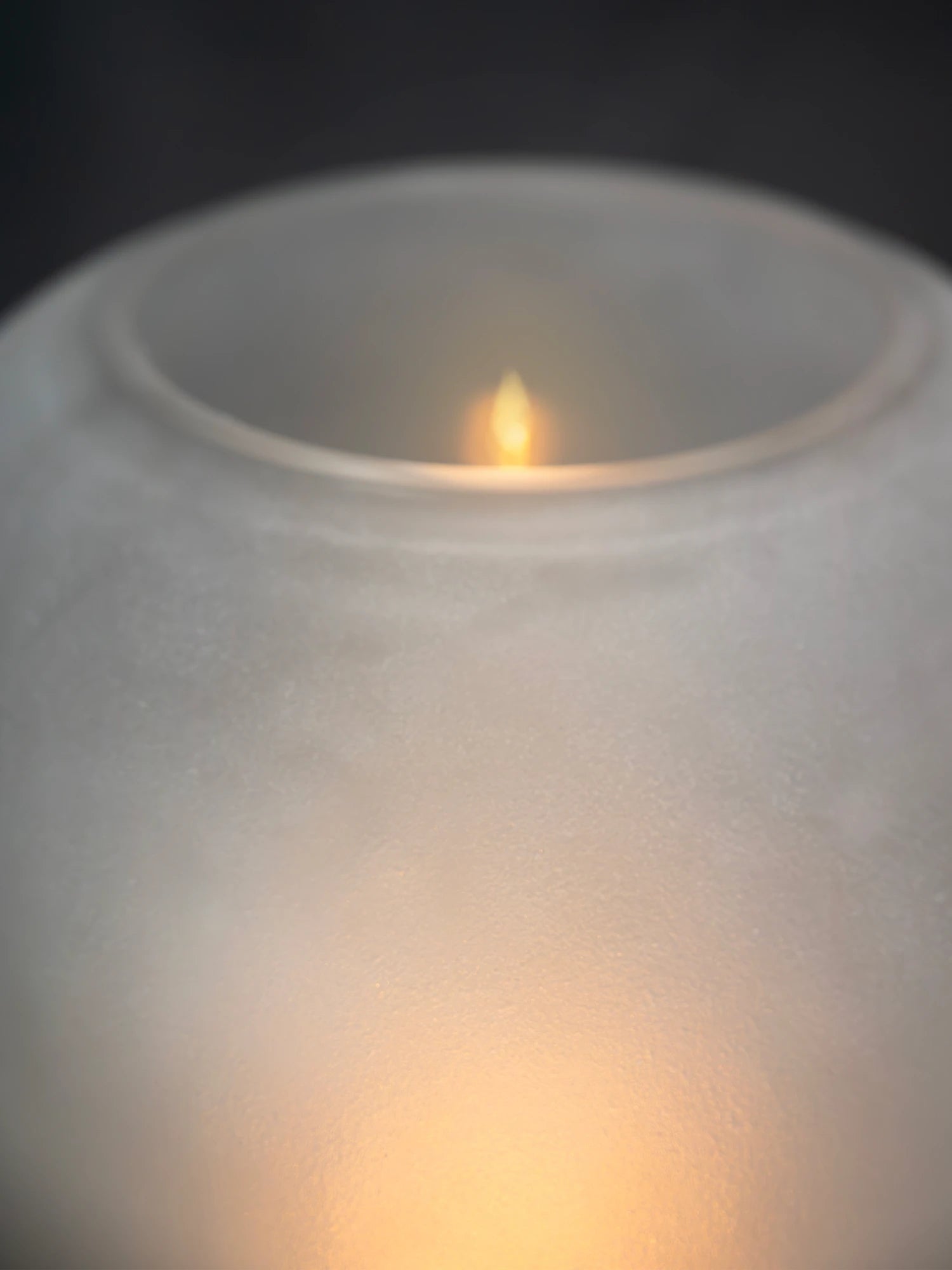 A high-angle close-up of the Lumen Wind Light's frosted glass globe, looking down into the opening. The soft, warm yellow-orange light is concentrated at the bottom, and a small, focused, flame-like glow is visible near the center of the opening, suggesting a candle or small bulb inside. The background is a dark gray/black.