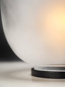 A dramatic close-up, low-angle shot of the Lumen Wind Light. The image focuses on the lower curve of the frosted, matte glass globe where it meets the thin, textured black base. A bright, warm yellow-orange light is visible from the right side, illuminating the interior of the globe. The background on the left is a dark shadow.
