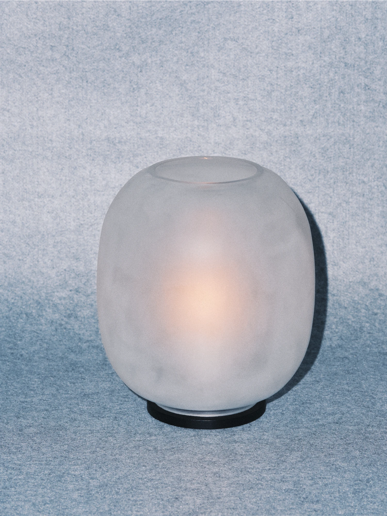 A Lumen Wind Light featuring a smooth, frosted, matte glass globe with a soft, warm glow emanating from within. The globe rests on a thin black circular base. The light is photographed against a background of mottled, textured blue-gray fabric or paper.