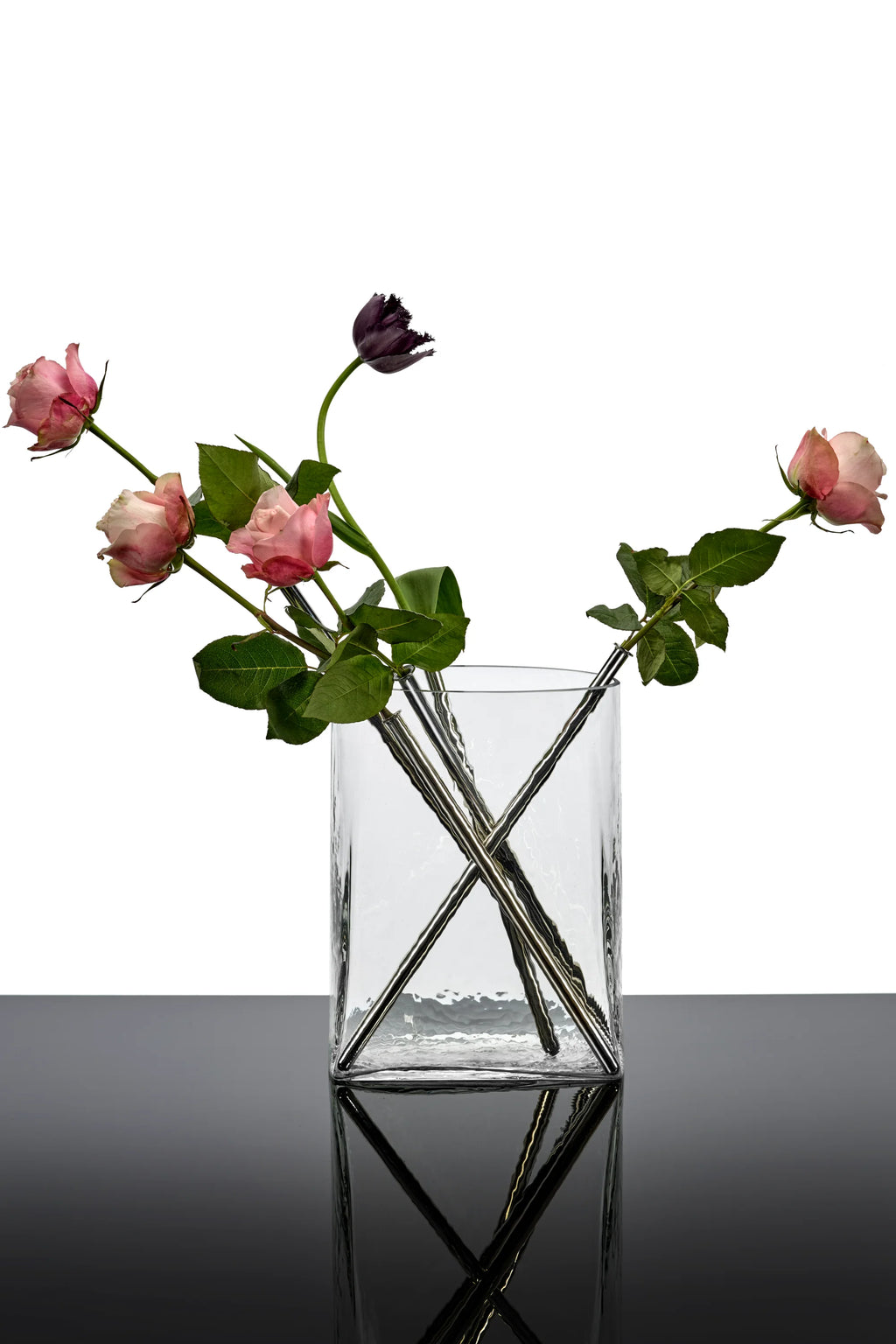 Serax Josephine Vase with Glass Tube 03, a clear, rectangular mouth-blown glass vase featuring a textured, hammered finish and four integrated silver-toned glass tubes arranged in an "X" pattern, holding pink roses and a dark purple tulip. This elegant decorative vase is perfect for modern home decor enthusiasts seeking a unique floral display.