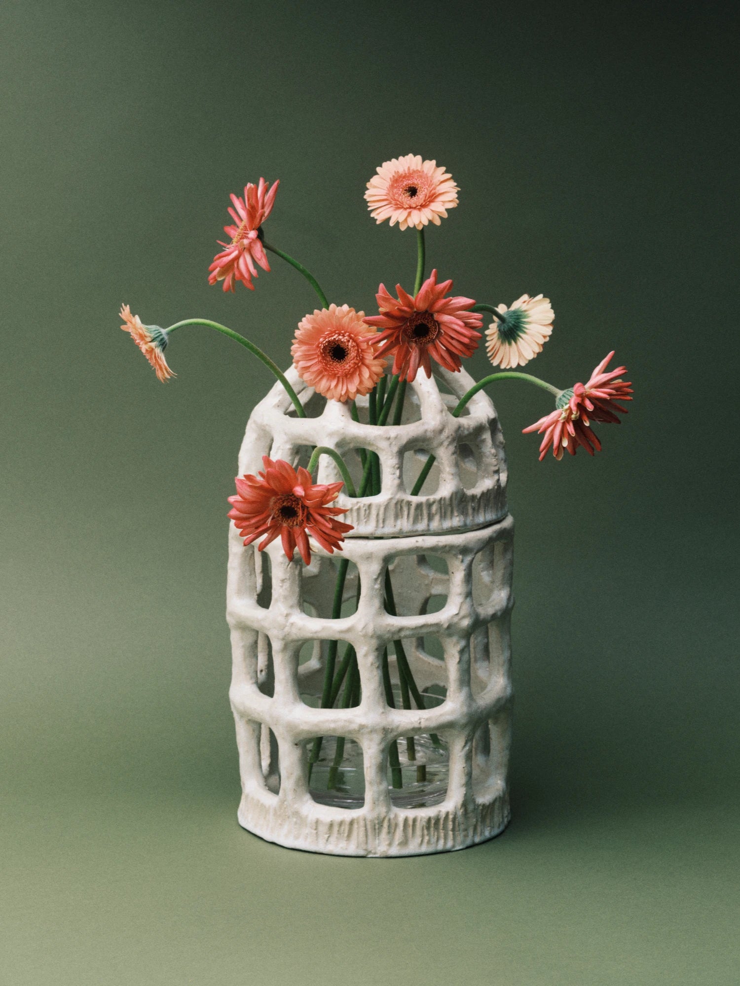 A white, textured ceramic Pique-Fleurs Vase featuring a decorative, cage-like pierced design with rectangular and oval cutouts. The two-piece vase contains several orange and coral-colored gerbera daisies with long, curving stems, which are held in place by the perforated structure. The vase is set against a solid, dark, olive-green background.