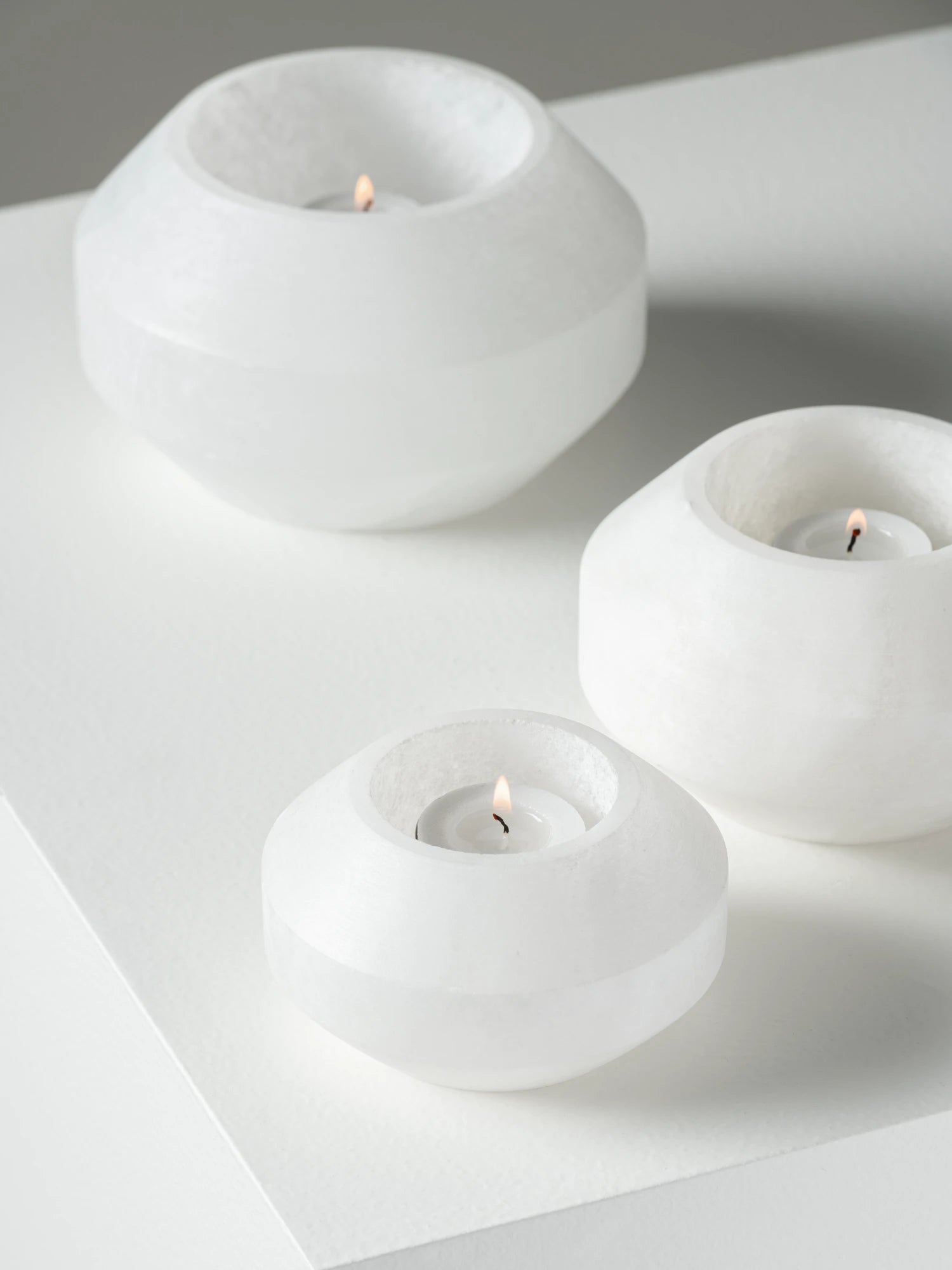 A high-angle studio shot of three faceted alabaster tealight holders of different sizes arranged on a white surface. All three holders are made of translucent white alabaster and contain small, lit tealight candles with visible flames. The holders are rounded but feature horizontal facets, creating a subtle contrast against the bright, minimalist background.