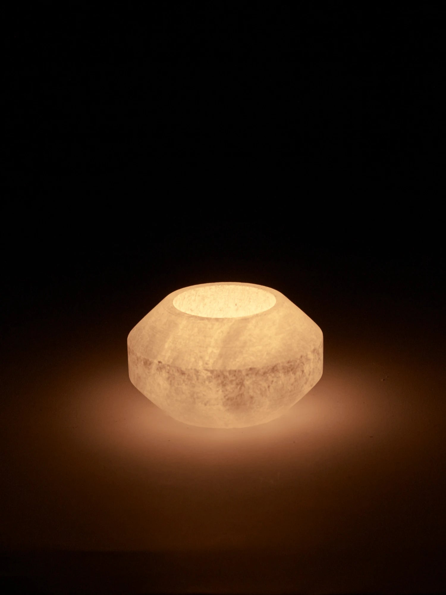A small, low, faceted alabaster tealight holder in a studio shot. The holder is lit from within, emitting a soft, warm orange and yellow glow that highlights the natural translucence and veining of the stone. The glow illuminates the surface around it, while the background is completely black.