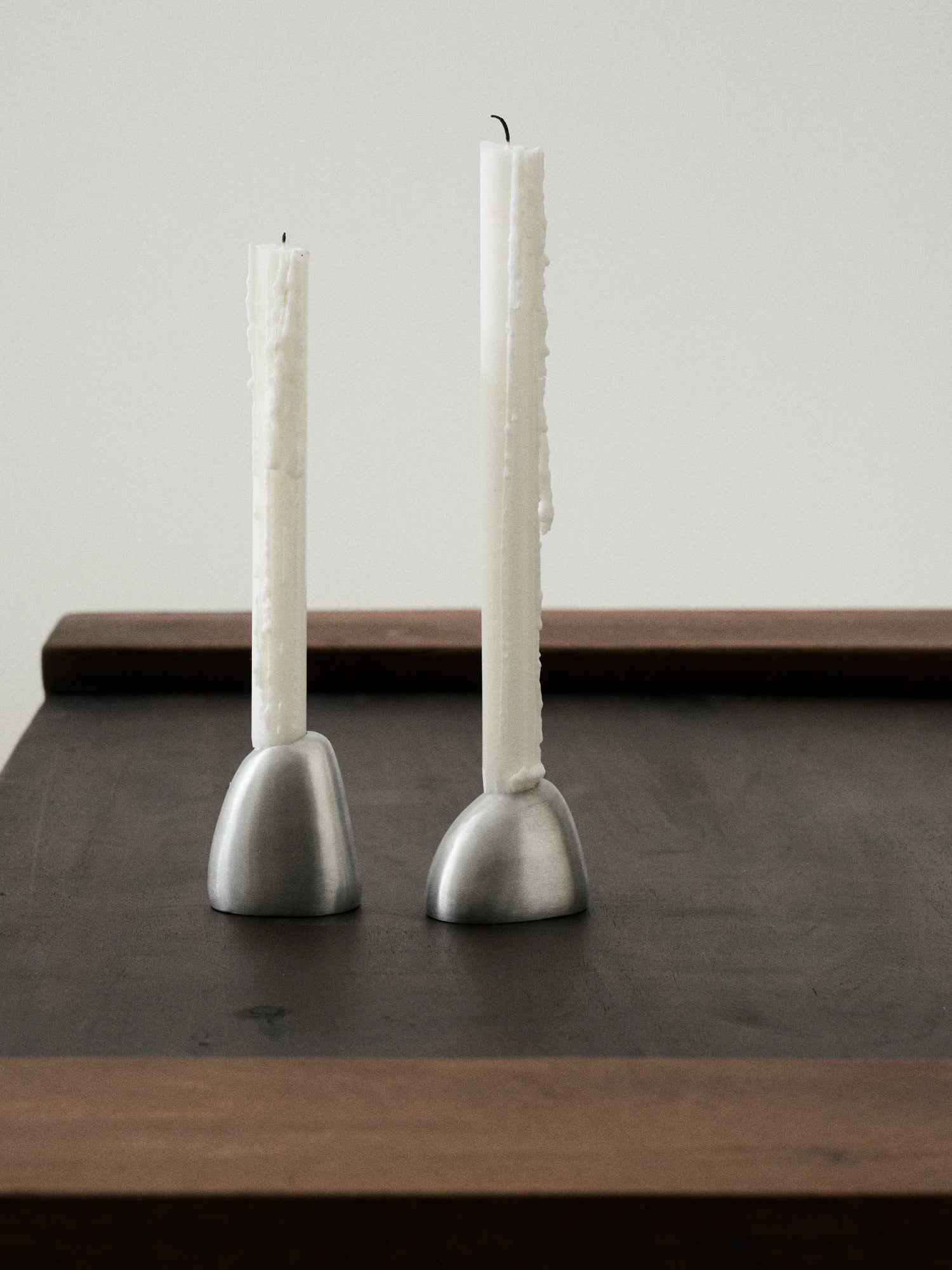 A lifestyle close-up showing two Out of Lines Candle Holders made of brushed silver or pewter metal with a smooth, domed base that subtly tapers upward. Each holder is topped with a partially burned, thick white pillar candle that has visible drips of wax running down its sides. The holders are placed on a dark wood surface with a warm brown frame against a plain white or light gray background.