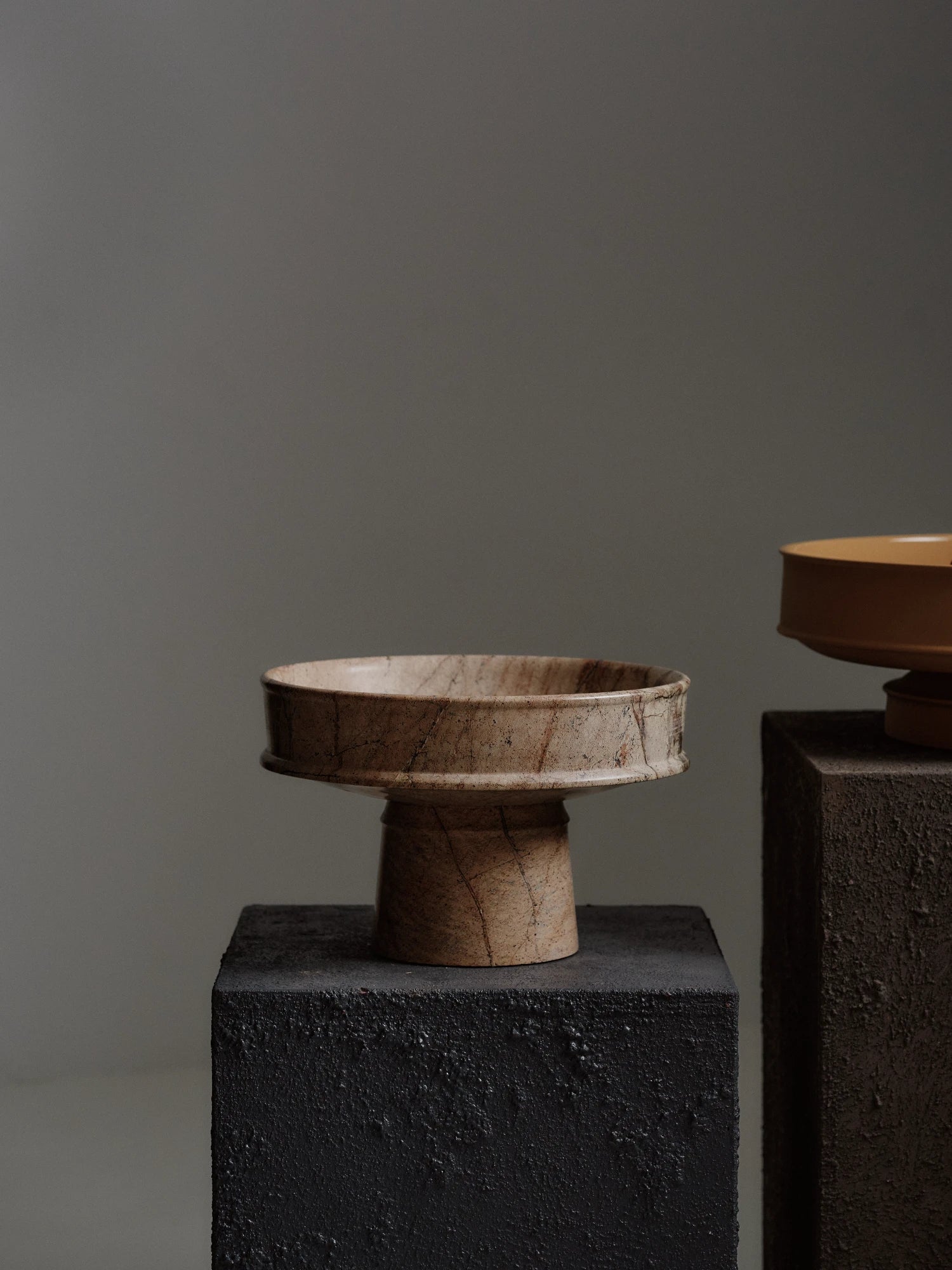 The Dune Raised Bowl made of light brown, veined marble is shown on a dark gray textured plinth on the left. To the right, a smooth orange/terracotta ceramic raised bowl holds a small bunch of red grapes. The background is a solid dark gray.