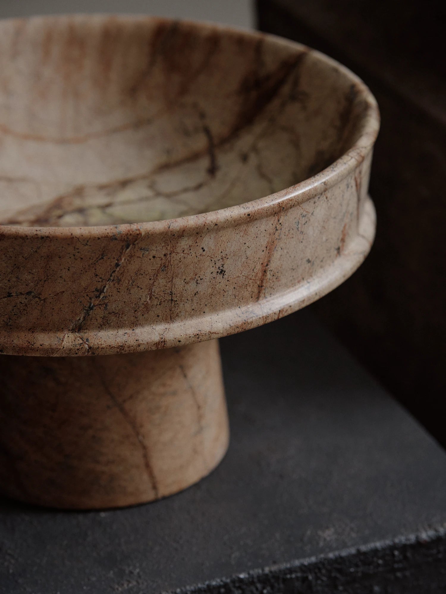 An extreme close-up of the Dune Raised Bowl made of light brown, finely cracked and veined marble. The focus is on the bowl's rounded rim and the cylindrical pedestal base as they rest on a dark, rough-textured surface.