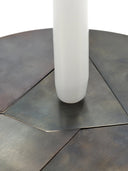 A close-up shot of the Candelabra Blue Steel Candle Holder's round base, showing the brushed, dark steel surface composed of geometric, triangular segments pieced together. A section of a thick, white wax candle is visible vertically in the center right, inserted into the base. The background is white.