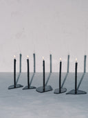 Five individual Candelabra Blue Steel Candle Holders are lined up diagonally across a light gray surface. Each holder features a single slim, dark-colored candle fixed to a small, geometric dark steel base. All five candles are lit with small flames, casting long, dark, vertical shadows onto the light gray wall behind them.