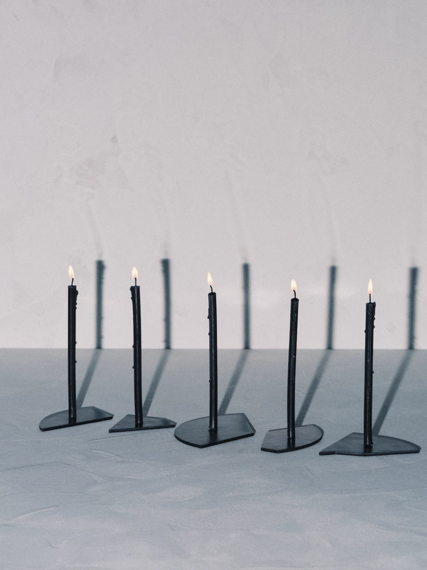 Five individual Candelabra Blue Steel Candle Holders are lined up diagonally across a light gray surface. Each holder features a single slim, dark-colored candle fixed to a small, geometric dark steel base. All five candles are lit with small flames, casting long, dark, vertical shadows onto the light gray wall behind them.