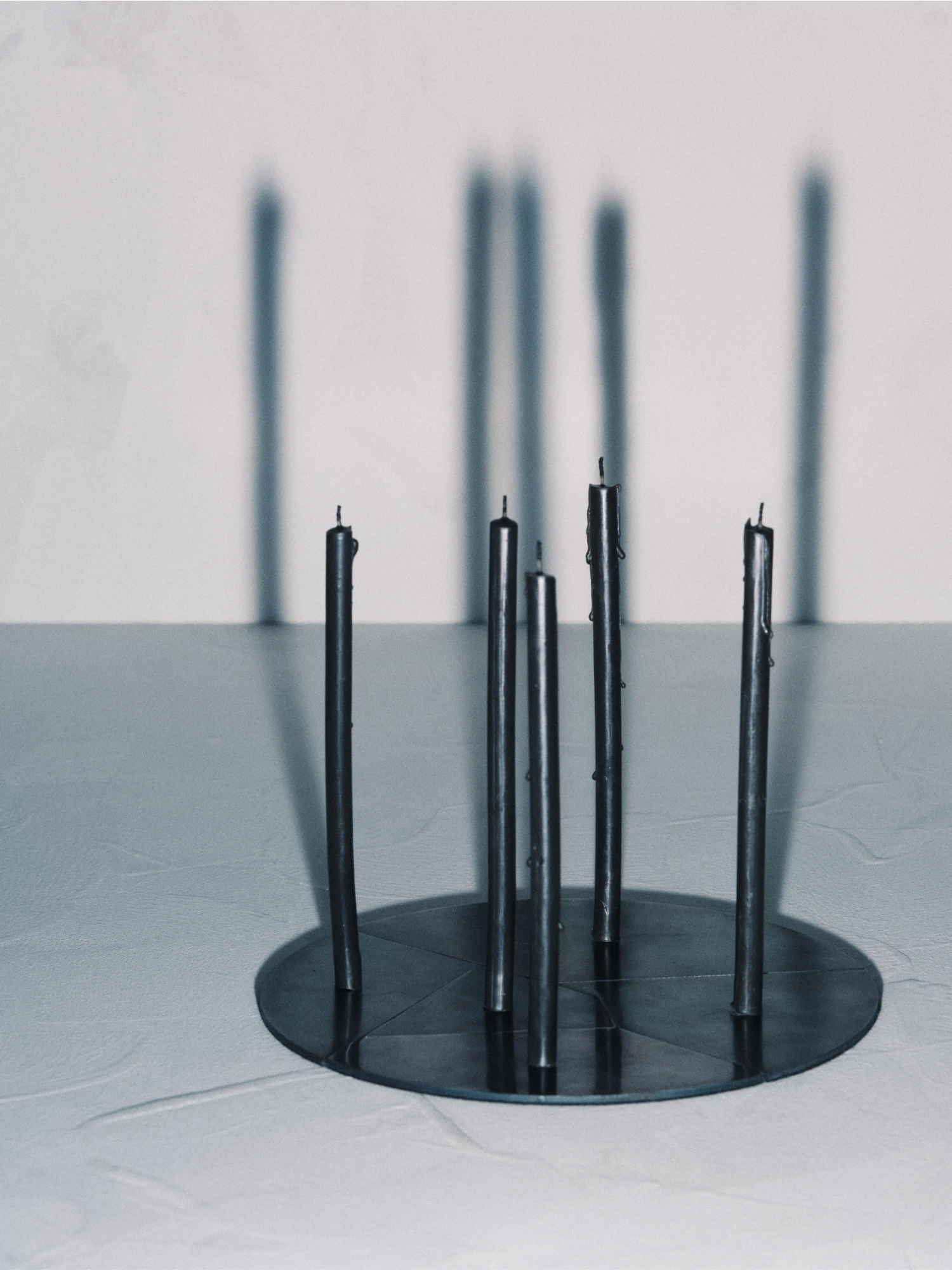 The Candelabra Blue Steel Candle Holder with a round, dark steel base holds five tall, slim, dark-colored candles. The candles are unlit, showing their charred wicks, and are casting long, vertical shadows onto the light background wall. The holder and candles are placed on a light-colored surface.