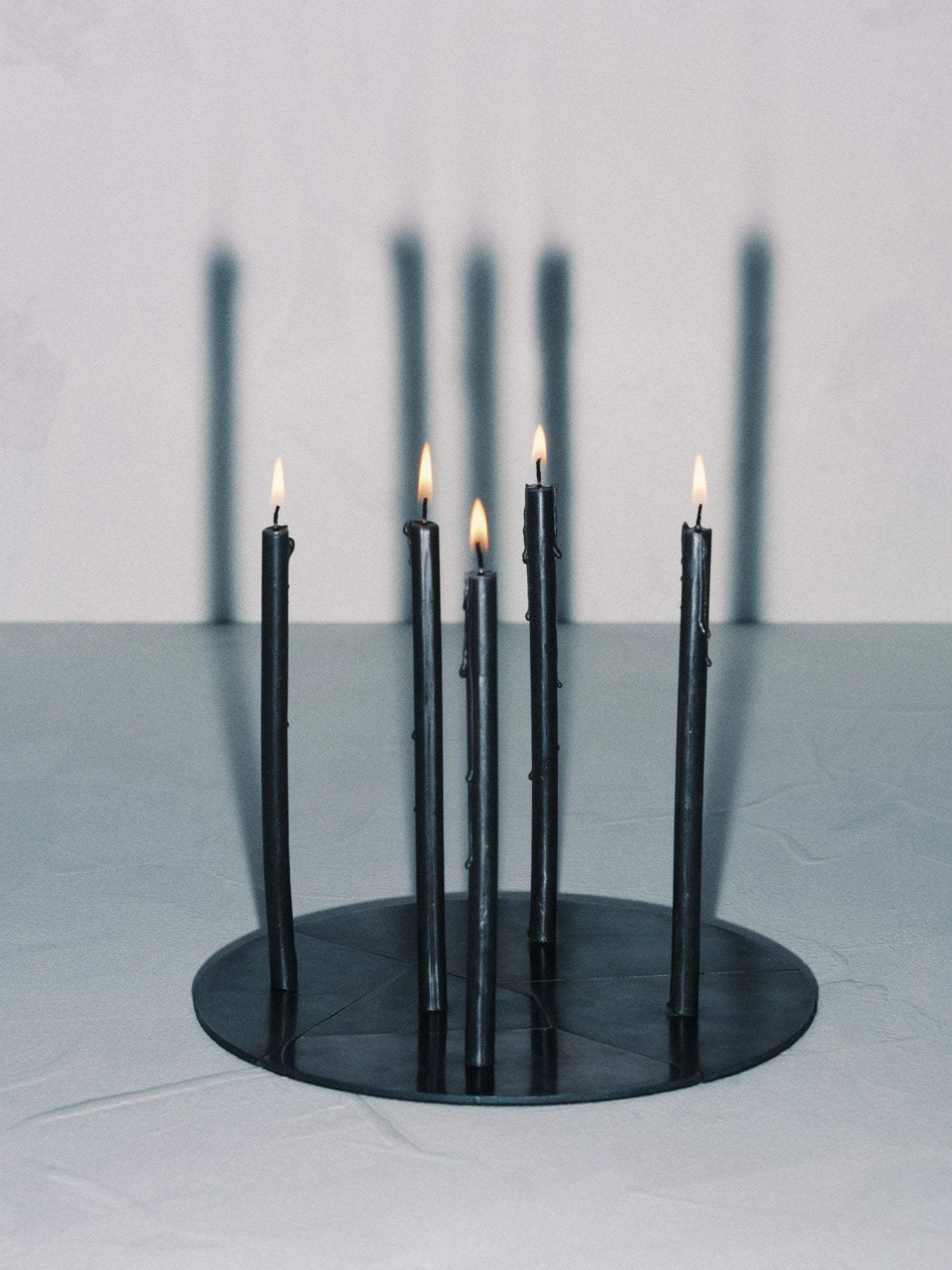 The Candelabra Blue Steel Candle Holder with a round, dark steel base holds five tall, slim, dark-colored candles that are currently lit, with small flames at the top. The candles and base are placed on a light-colored surface, and their long, vertical shadows are cast dramatically on the light background wall.