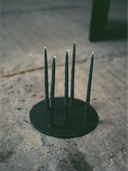 A low-angle shot of the Candelabra Blue Steel Candle Holder, featuring a round, dark steel base with five vertical thin, dark-colored candles inserted into the top. The candles are unlit, showing their white wicks. The candle holder is placed on a rough, textured, gray concrete or stone surface.