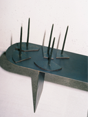 A conceptual display featuring five individual Candelabra Blue Steel Candle Holders—each with a slim, dark-colored taper candle inserted into a small, geometric dark steel base—arranged on a larger, elongated, dark blue-black steel platform or table. The platform has rounded ends and is supported by a single vertical steel leg. The unlit candles cast shadows onto the light gray wall behind them.
