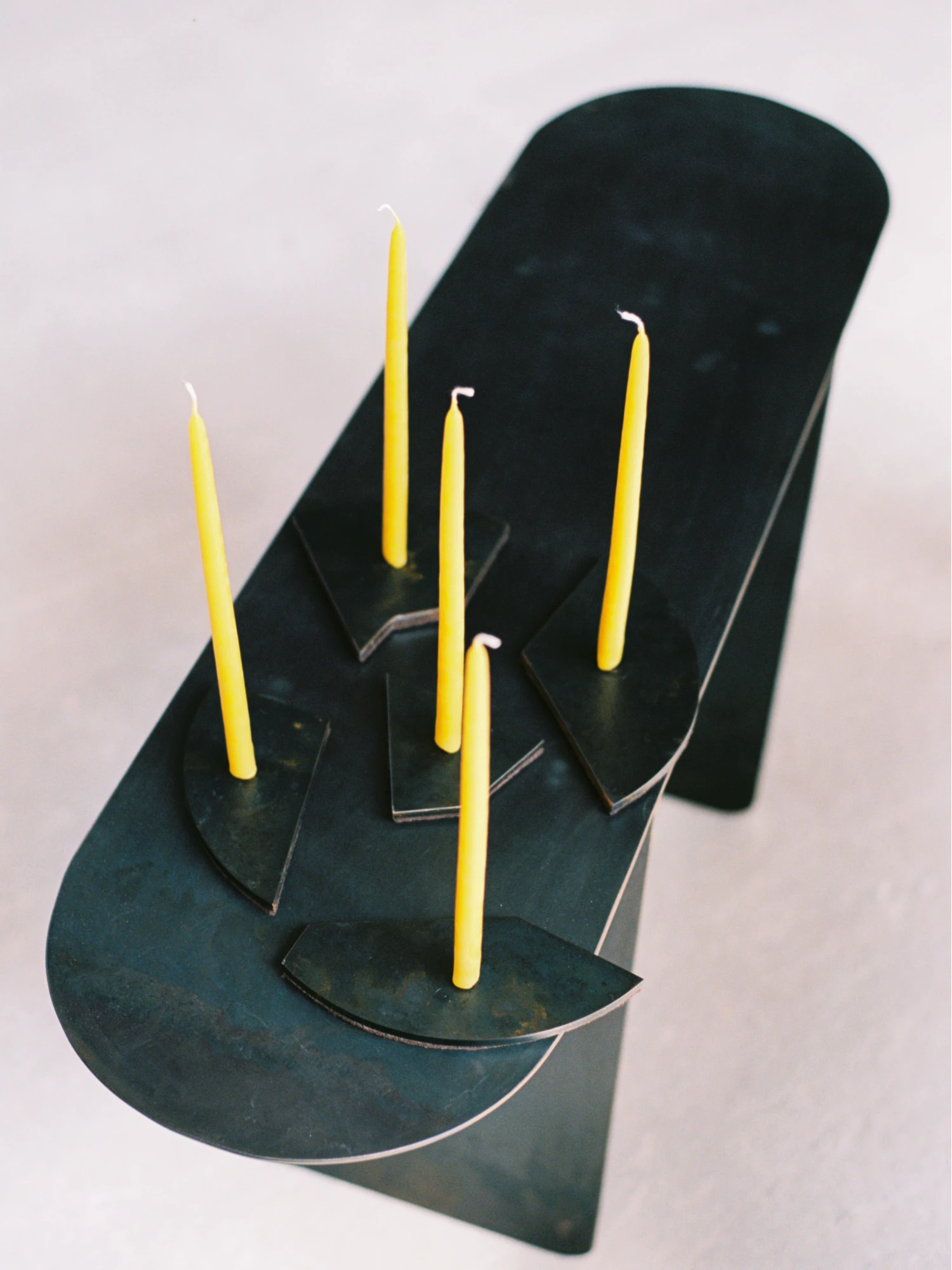 A high-angle close-up of the Candelabra Blue Steel Candle Holder displayed in a unique, layered arrangement. Five individual dark blue-black steel bases, each holding a bright yellow taper candle, are arranged on top of a larger, curved, dark blue-black steel platform or sculptural element that stands vertically in the background. The candles are unlit, and the overall color palette is dark blue and vibrant yellow against a light background.