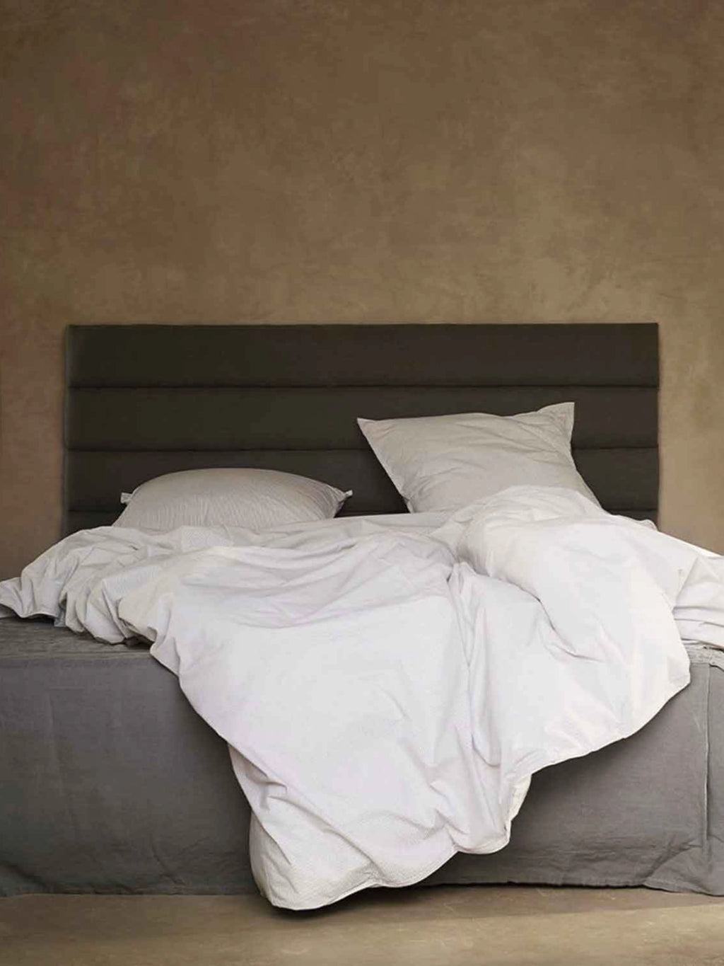 Tine K Linen Headboard, a modern minimalist upholstered headboard in dark charcoal grey linen featuring distinct horizontal paneled detailing. This soft, inviting piece provides a serene backdrop, complementing the relaxed white linen bedding and grey bed skirt for a cozy Scandinavian bedroom aesthetic. Ideal for creating a calming and contemporary bedroom design.