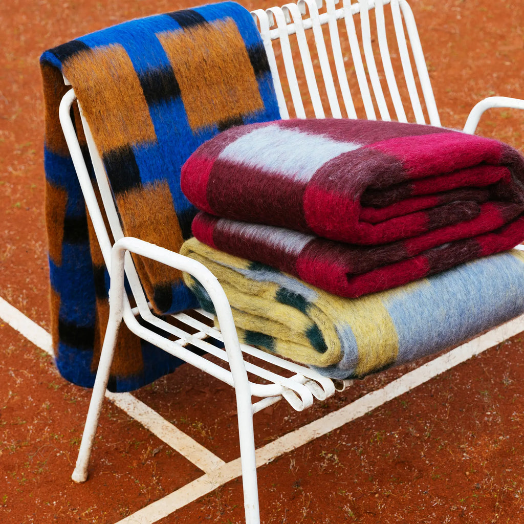 Layered Dancing Doll Oversized Throw Blanket, showcasing three plush, colorful patterned throws draped over a white slatted bench on a red tennis court. Perfect for adding cozy style and warmth to...