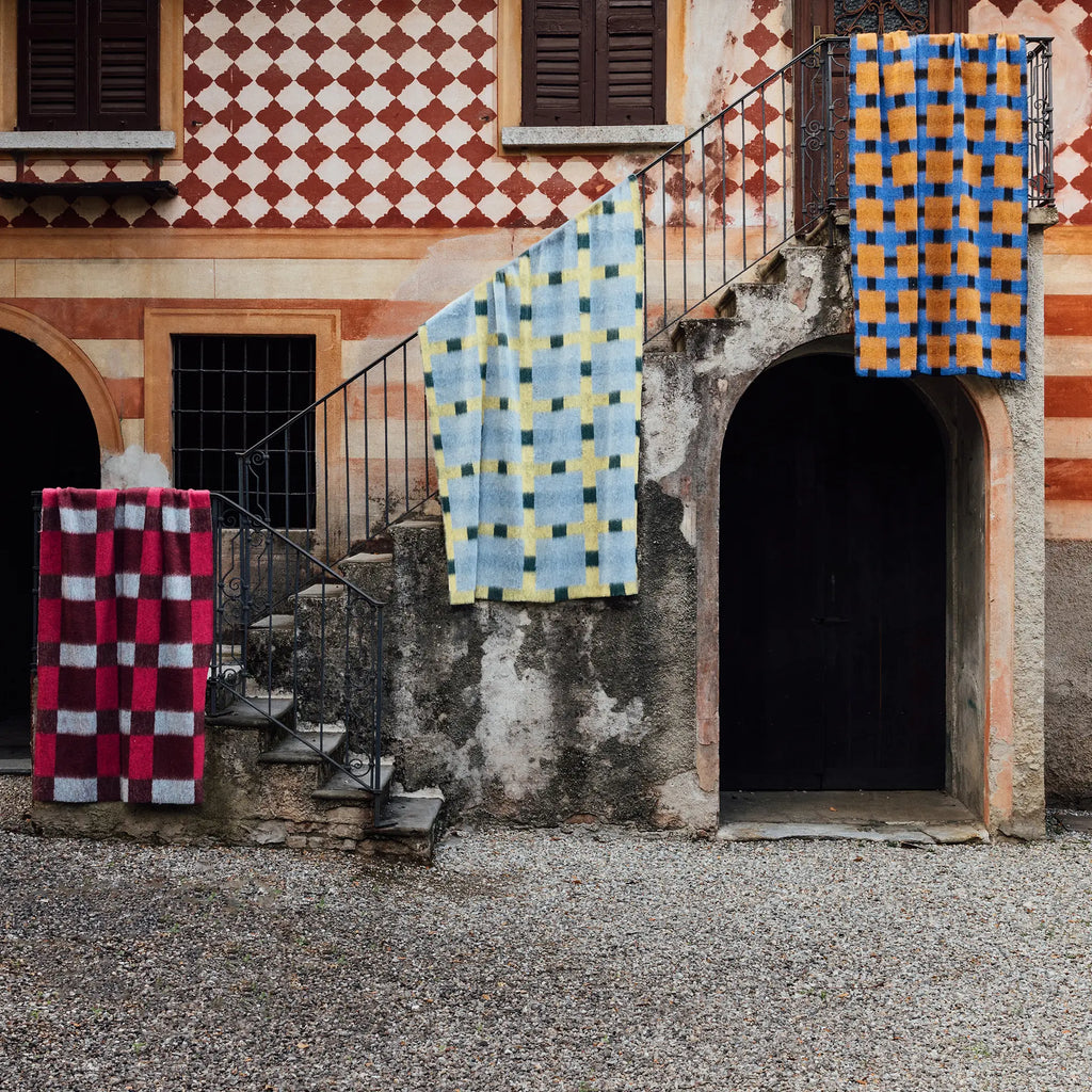 Layered Dancing Doll Oversized Throw Blanket collection, featuring three checkered blankets in blue/orange, light blue/yellow, and red/burgundy patterns, draped on a rustic stone staircase of an ol...