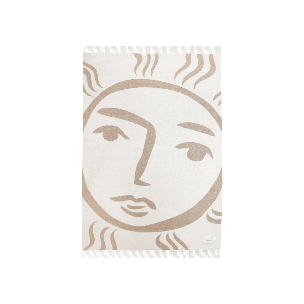 Layered Leggero Throw Blanket, a soft cream-colored throw with a bold beige abstract sun face design and delicate fringe edges. Perfect as a decorative and cozy accent for living rooms or bedrooms.