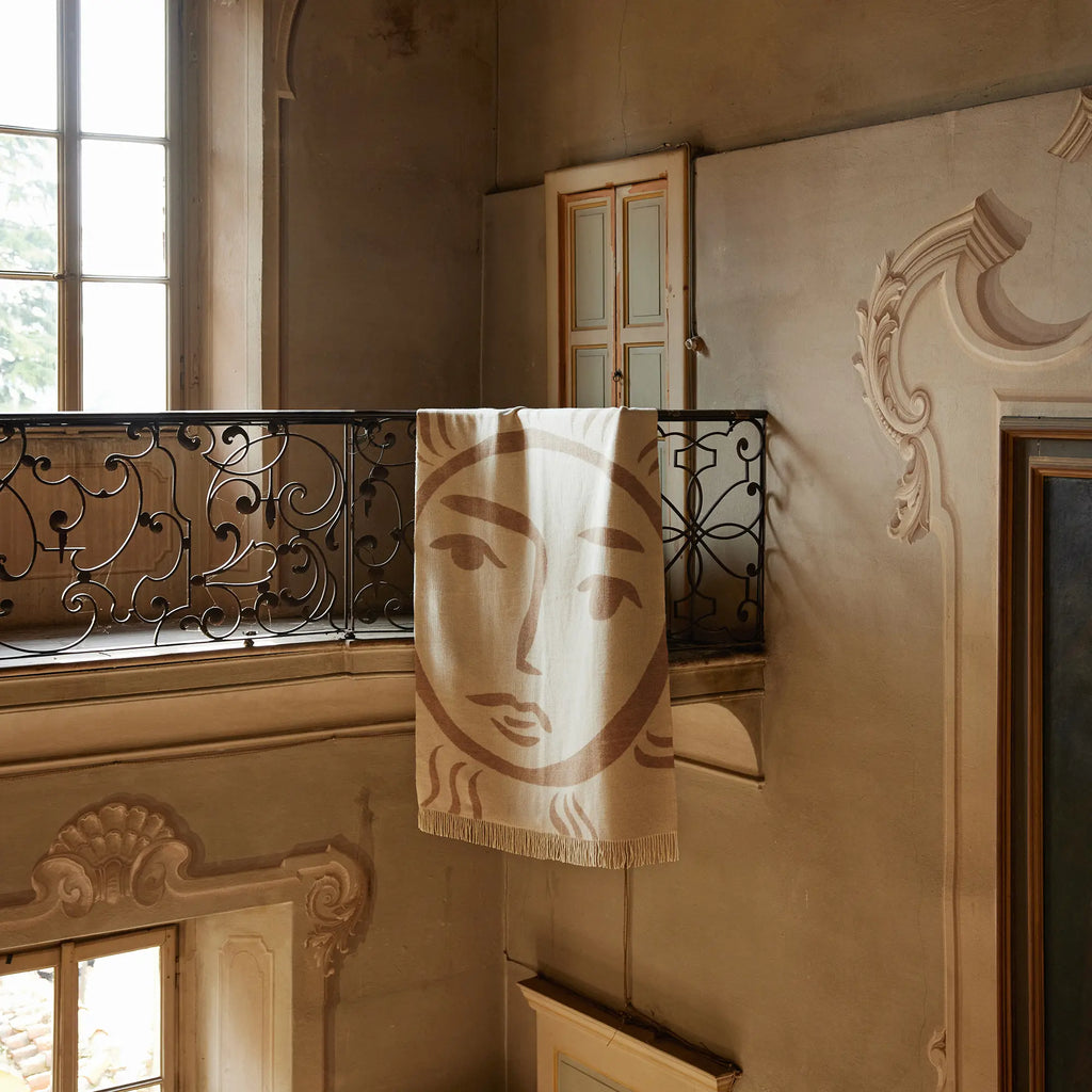 Layered Leggero Throw Blanket, a cream and brown patterned throw with a sun face design and fringed edges, draped over a decorative wrought iron railing in an old, ornate building. Perfect for addi...