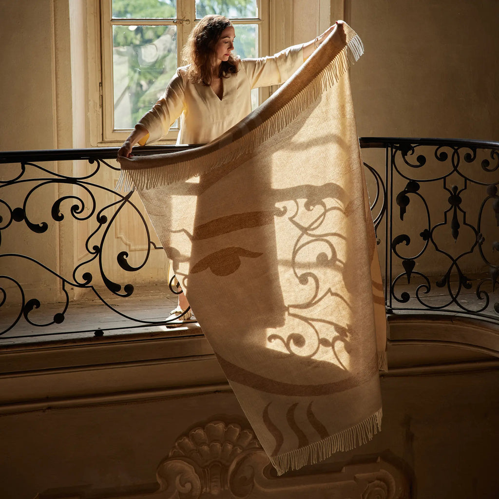 Layered Leggero Throw Blanket, a light beige and brown patterned throw with fringe edges, held by a woman on a balcony. Perfect for adding stylish comfort and warmth to any living space.