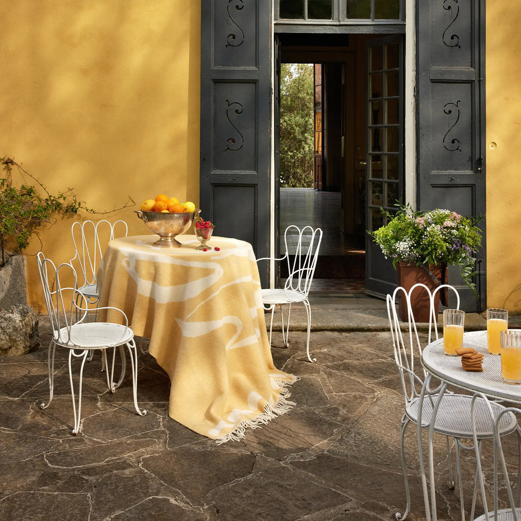 Layered Fruttato Throw Blanket, a soft yellow wool throw with white abstract wavy patterns and fringed edges, draped over a round outdoor table. Perfect for adding warmth and style to outdoor livin...