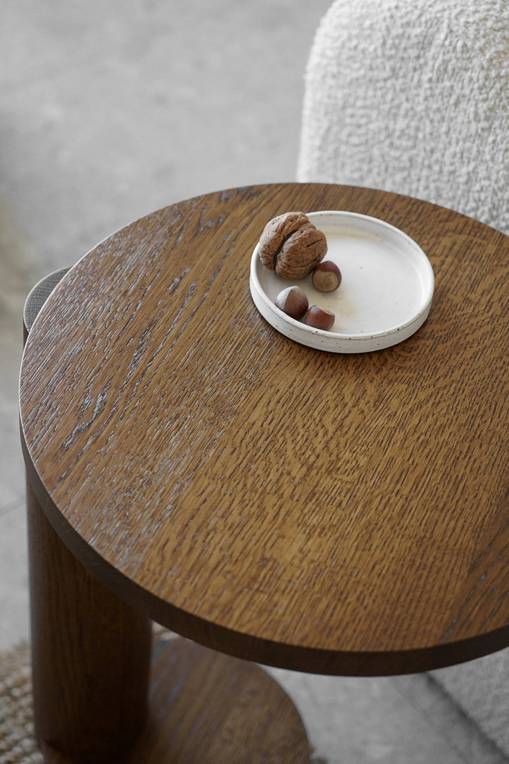 TAMO Bold Side Table, showcasing its dark brown, rich wooden texture and round top with a sturdy cylindrical leg. A white ceramic dish with a walnut and hazelnuts is placed on top, next to a light-colored, fluffy armchair, highlighting its premium design. Ideal for modern living spaces.