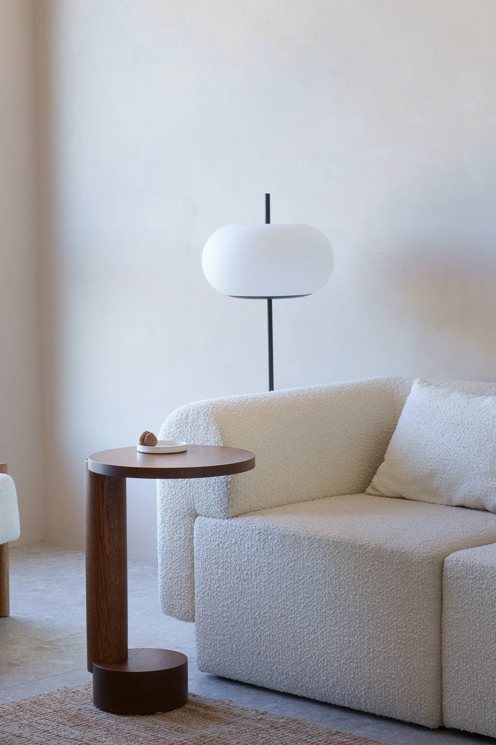 TAMO Bold Side Table in a rich dark brown, showing its circular top resting on a thick cylindrical pillar and round base, styled next to a cream boucle sofa. This premium wooden accent is perfect for modern living spaces.