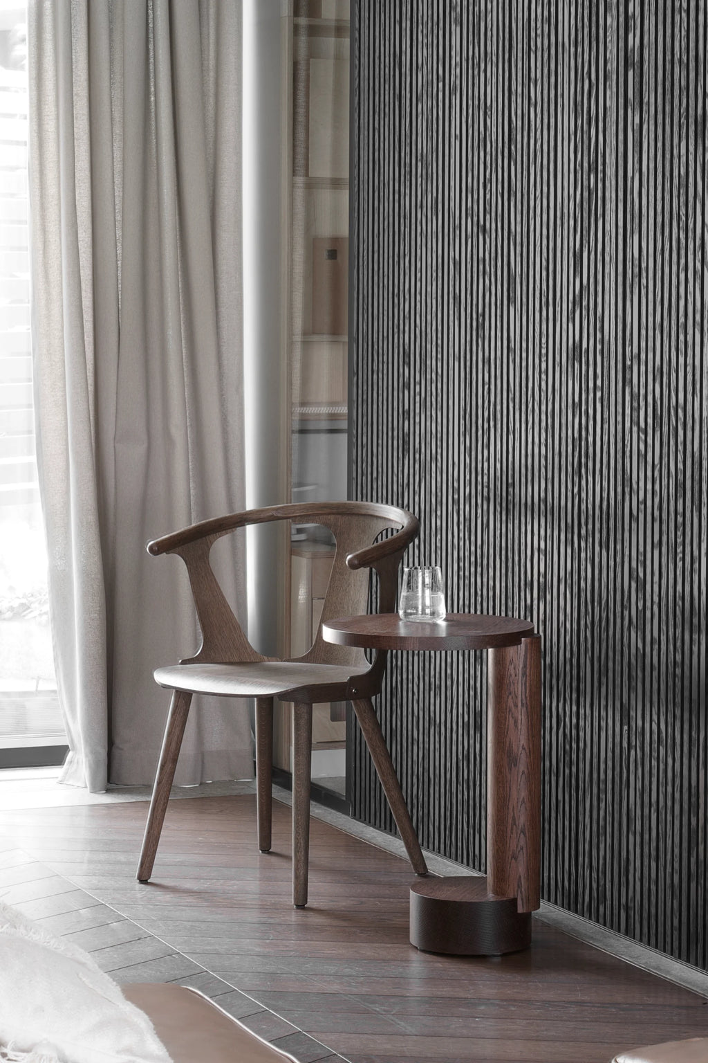 TAMO Bold Side Table, displayed next to a wooden chair with a clear glass of water on its round dark brown top, showcasing its handcrafted wood grain. Perfect for contemporary living spaces.