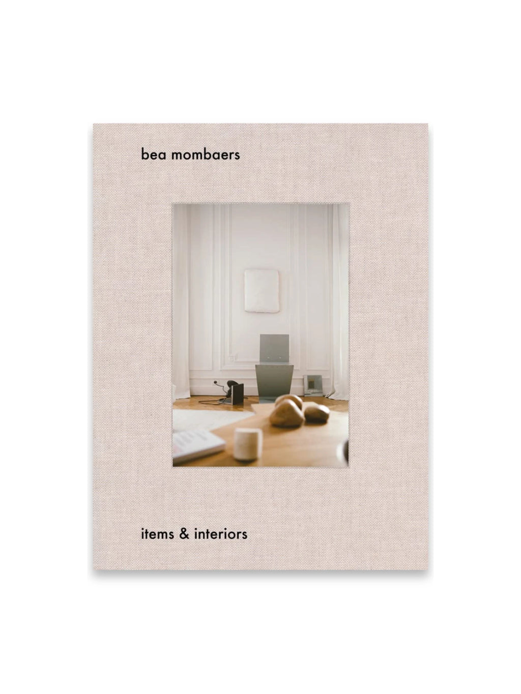 Book Bea Mombaers – items & interiors, a premium hardcover book with a textured, light beige linen-like cover. The front features a central image of a stylish interior with a laptop, natural stones, and a ceramic cup on a wooden table. This elegant coffee table book is perfect for interior design enthusiasts and lifestyle readers seeking inspiration.