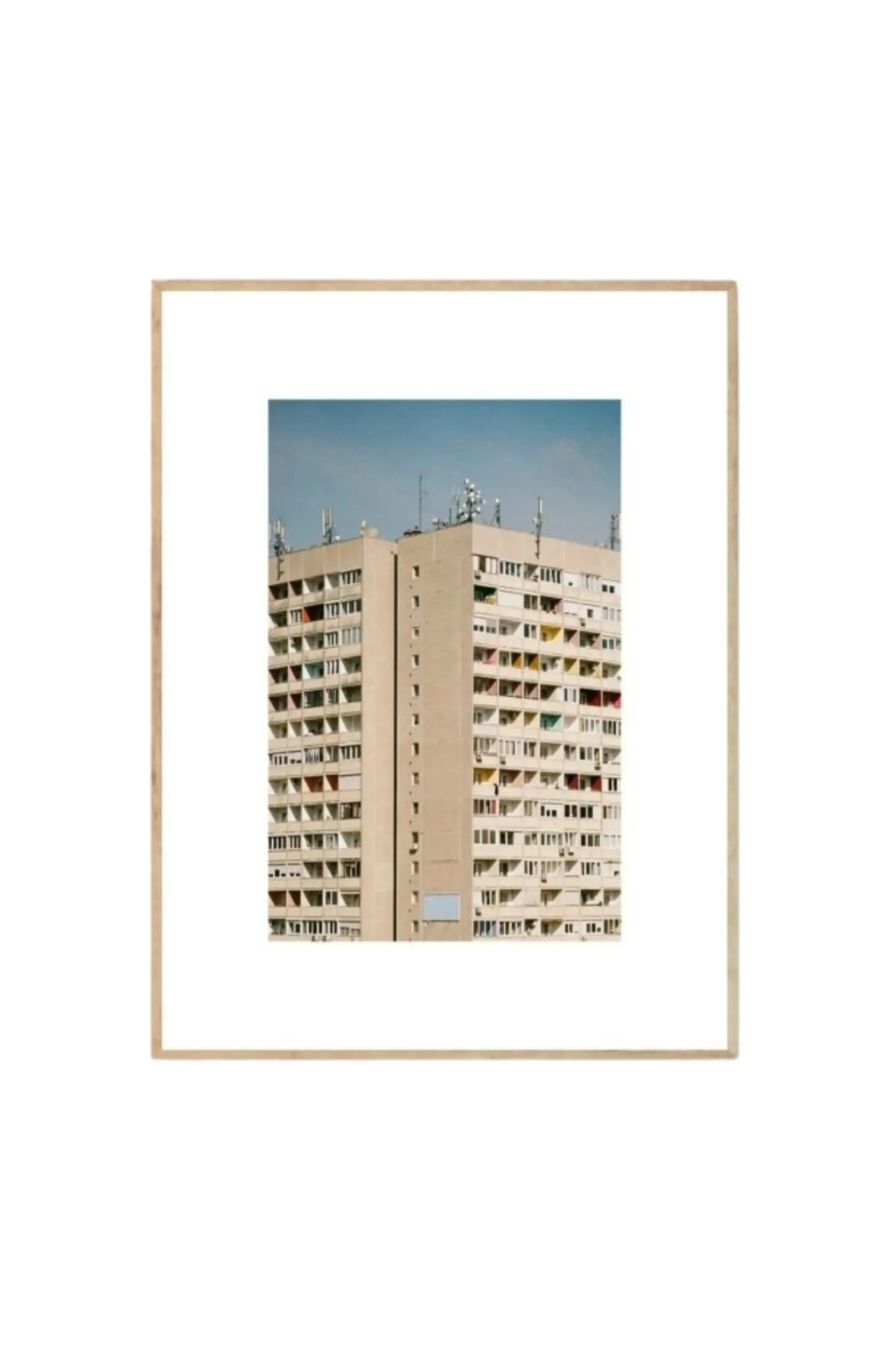 The Poster Club Block O’Clock - Art Print, a striking architectural poster featuring a tall, beige concrete building with numerous windows and balconies under a clear blue sky, framed in natural light wood. This unique photographic art print is perfect for modern urban dwellers and design enthusiasts looking for stylish wall decor.