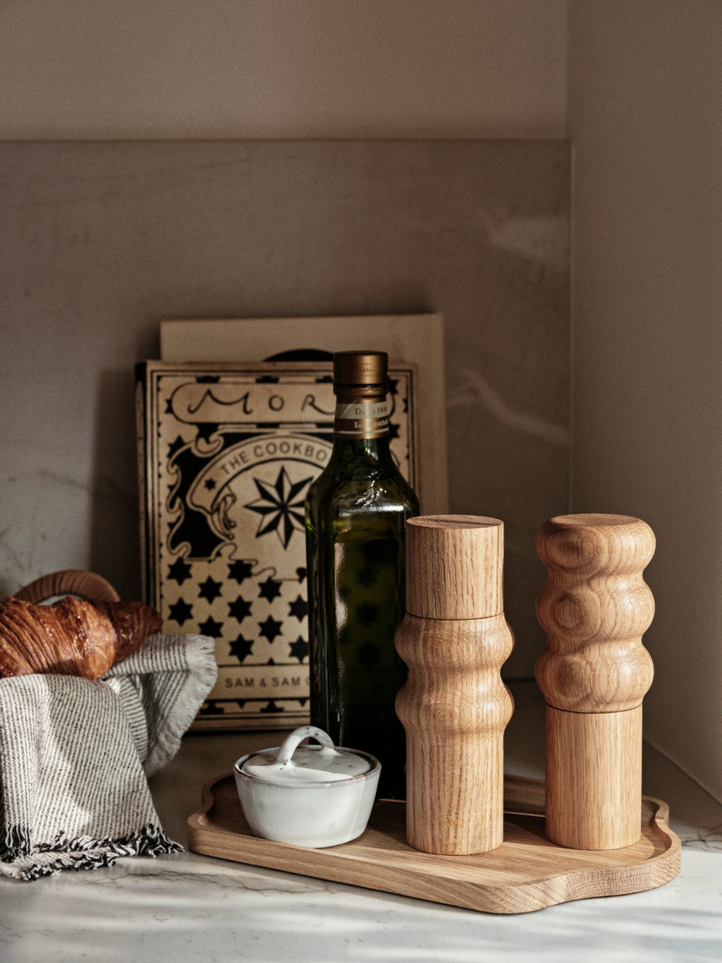 A pair of Edgar Salt & Pepper Mills made of natural wood with a sculptural, rounded, stacked-segment design, standing on a wooden tray. They are arranged next to a dark glass bottle of olive oil, a small white ceramic Nordic Sand bowl with a lid, and a cookbook. A croissant draped with a linen napkin is partially visible on the left.