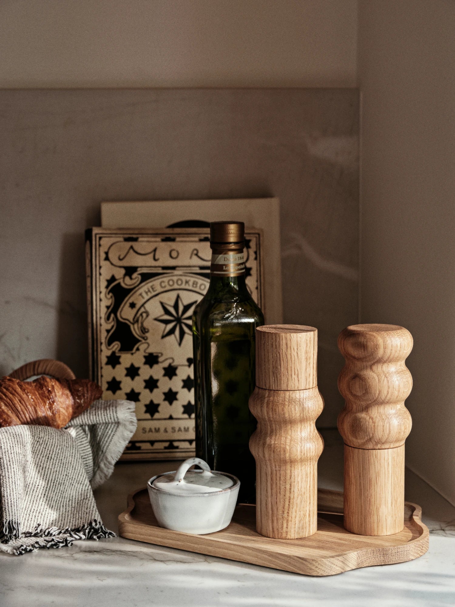 A pair of Edgar Salt & Pepper Mills made of natural wood with a sculptural, rounded, stacked-segment design, standing on a wooden tray. They are arranged next to a dark glass bottle of olive oil, a small white ceramic Nordic Sand bowl with a lid, and a cookbook. A croissant draped with a linen napkin is partially visible on the left.