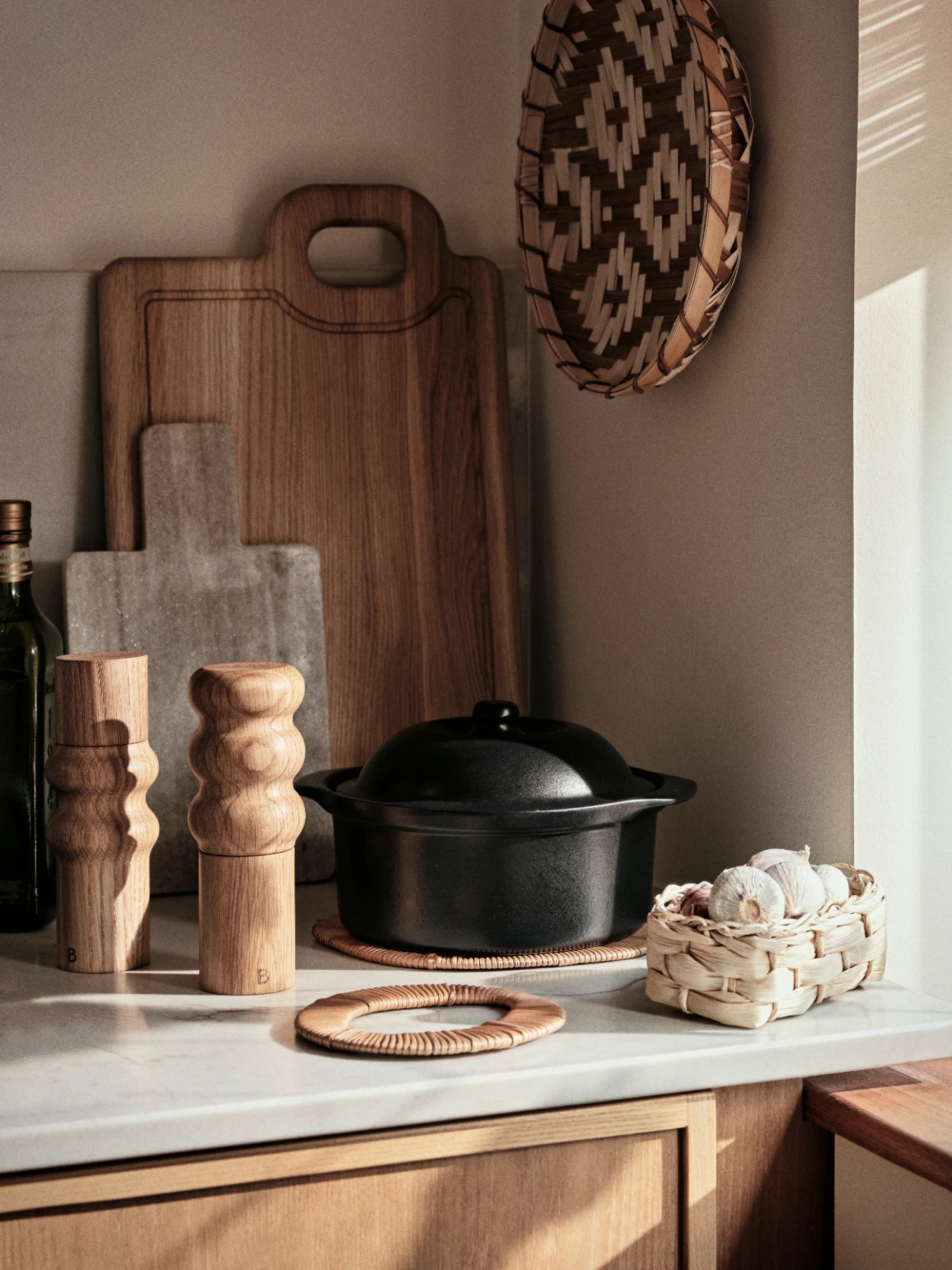 A pair of Edgar Salt & Pepper Mills with a natural wood grain finish and a distinct, rounded, stacked-segment shape, standing on a kitchen counter. They are positioned next to a black cast-iron Vig Ovenware pot sitting on a woven Tini Trivet, a small woven basket of garlic, a stack of wooden and marble cutting boards, and a hanging woven wall decoration.