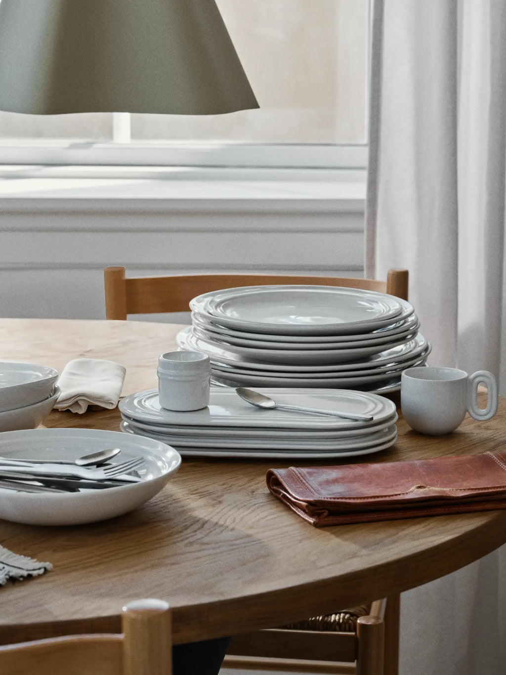 Broste Copenhagen Stevns Dinner Set for 4, a minimalist stoneware dinnerware set in a versatile light grey matte finish, featuring a rustic handcrafted texture and subtly raised rims. This contemporary Scandinavian tableware includes dinner plates, bowls, unique oval serving plates, and mugs, perfect for everyday dining or special occasions in modern minimalist and organic modern kitchens.