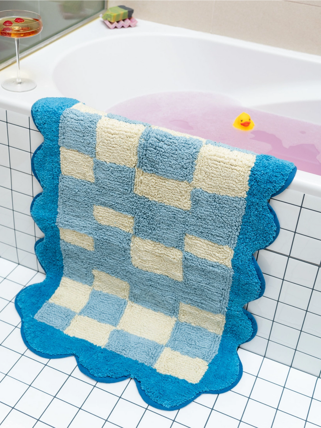 TARTA GELATINA Burbuja Bath Mat, a playful tufted cotton bath mat featuring a vibrant checkerboard pattern in bright blue, light blue, and cream hues, finished with a distinctive scalloped edge. This plush and soft bath rug adds a whimsical touch to contemporary bathrooms or eclectic powder rooms, perfect for buyers looking for fun bathroom decor.