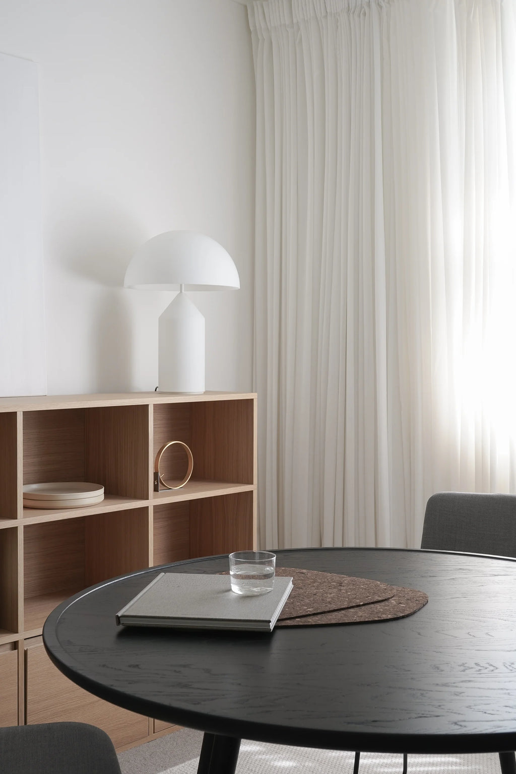 Mater Accent Round Dining Table, a dark stained oak finish, featuring a minimalist design with a smooth top and sturdy legs, styled with a book and glass of water, in a bright room with white curtains and a wooden shelving unit.