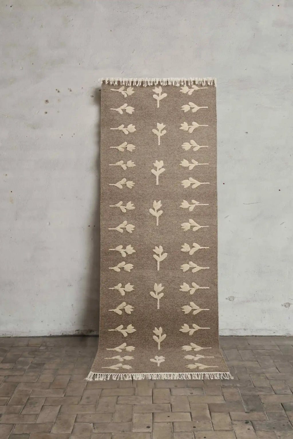 Cappelen Dimyr Colonnade No.09 Rug, a hand-knotted wool runner in a rich brown-taupe base color featuring raised cream botanical motifs of stylized leaves and flowers, with natural fringed ends. This organic modern rug offers a plush textured weave and a minimalist yet artistic design, perfect for adding warmth as a hallway runner, bedroom accent, or entryway rug in Scandinavian or boho-chic interiors.