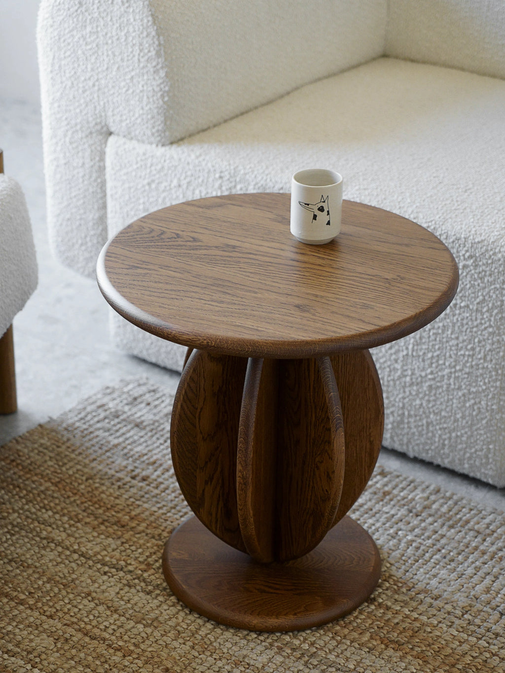 TAMO Citrus Side Table in a lifestyle shot. Features a round, rich dark brown oak top and a unique, sculptural slatted base, next to a cream boucle sofa and jute rug. A premium living room accent.