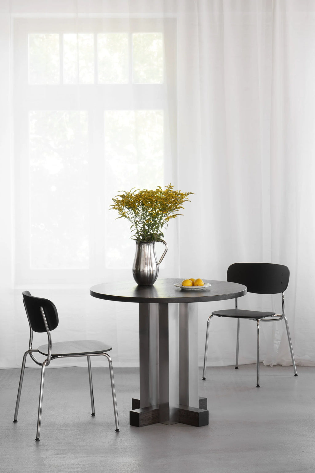 TAMO Cruz Round Table in a lifestyle shot. This premium modern dining table features a dark gray wooden top and a unique brushed stainless steel base with dark wood accents. Ideal for contemporary dining spaces.
