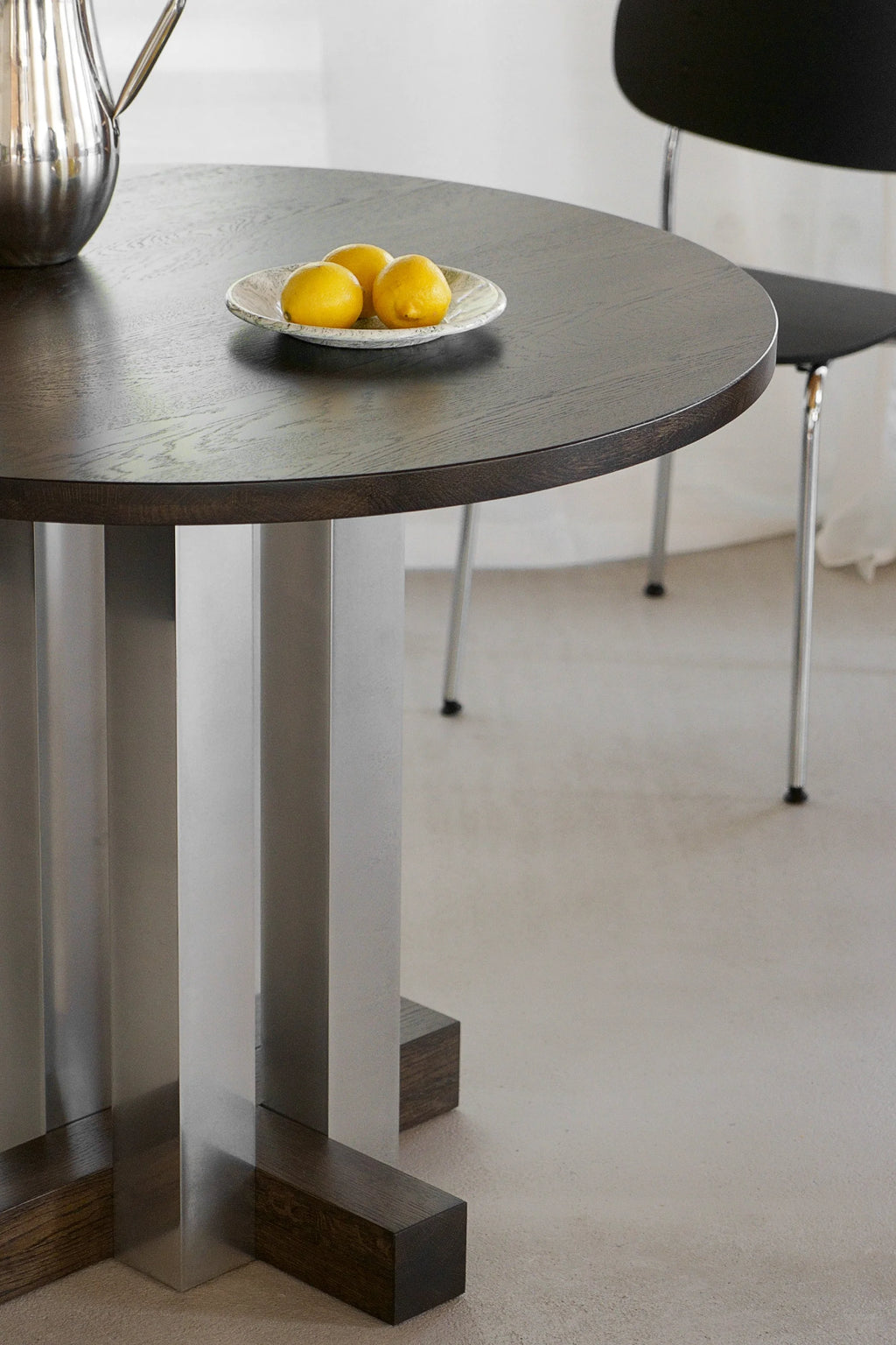 TAMO Cruz Round Table: Dark brown wooden round tabletop with visible grain, supported by a unique base of silver-gray rectangular pillars and dark wooden blocks. A premium furniture piece for any modern dining space.