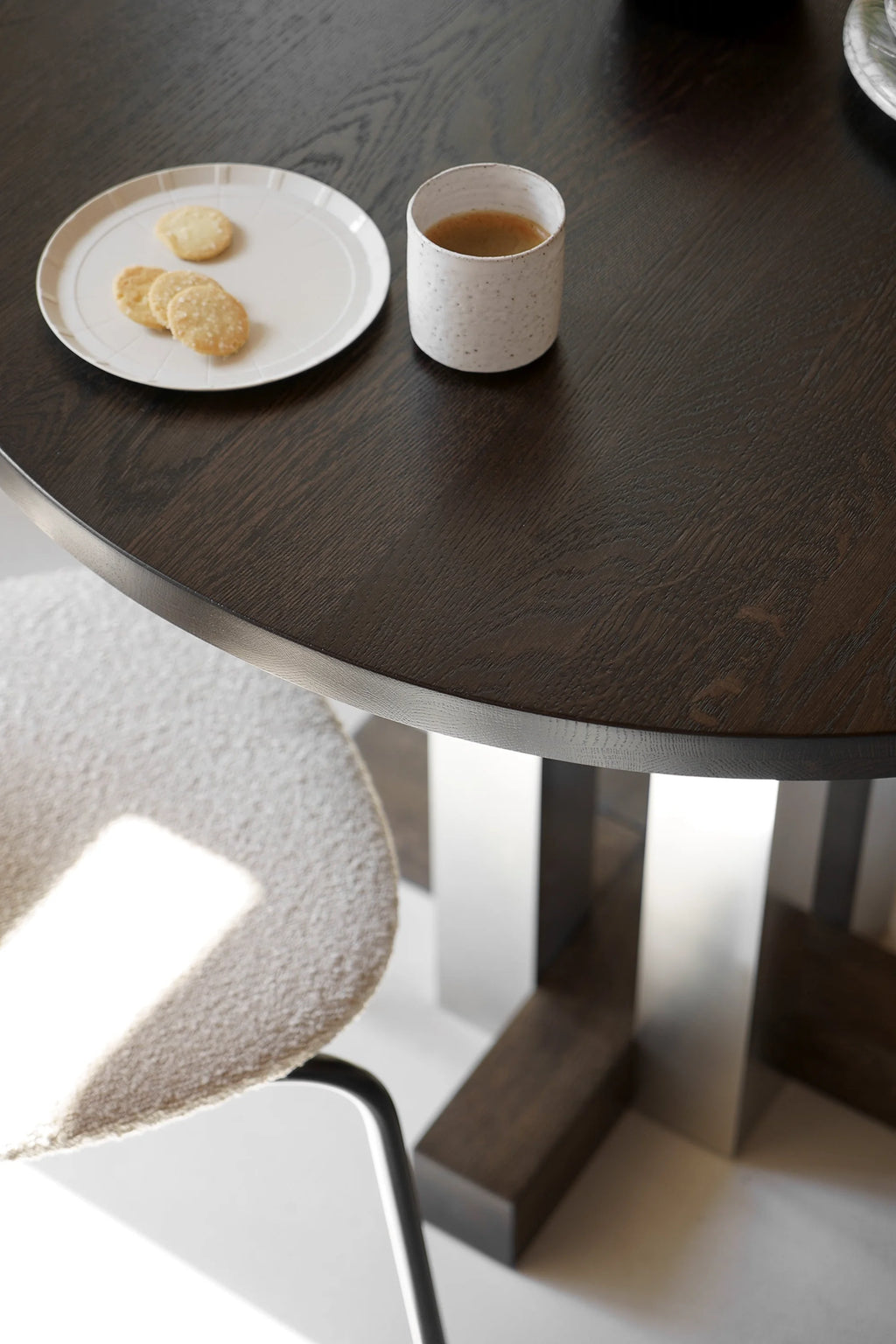 TAMO Cruz Round Table in a close-up, showcasing its dark brown wood grain surface with a beveled edge, paired with a light beige textured chair and contemporary base. A premium dining center.
