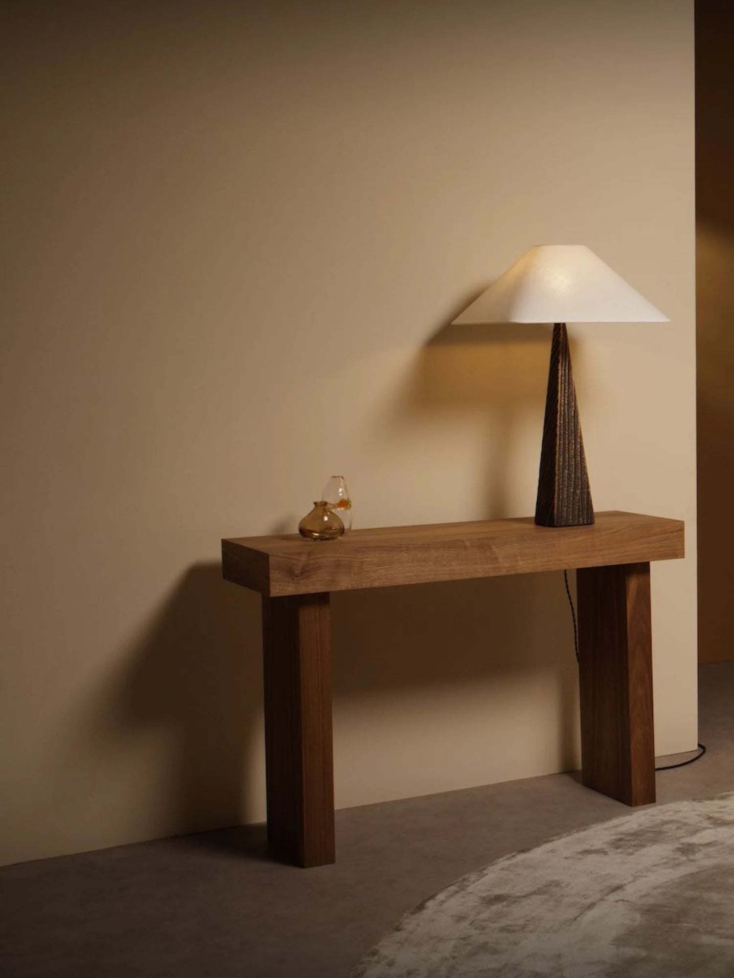 A Cubist Table Lamp with a textured, dark bronze or wood base and a wide, square, off-white lampshade is on a wooden console table. A small glass object is next to the lamp. The console table has a simple, minimalist design with two thick legs. A light-colored rug is partially visible in the bottom right corner.