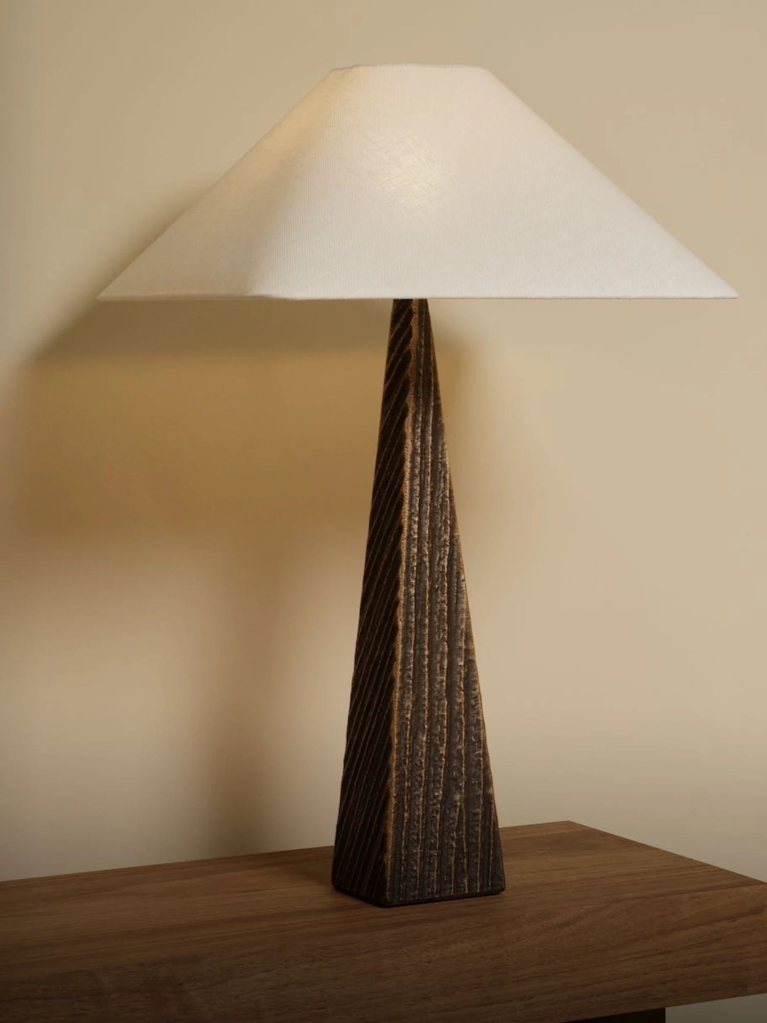 A minimalist Cubist Table Lamp is shown in a close-up shot, sitting on a wooden surface. The lamp has a tall, tapered, and twisted base with a rough, textured finish in a dark bronze or brown color. A wide, square, off-white lampshade is on top.